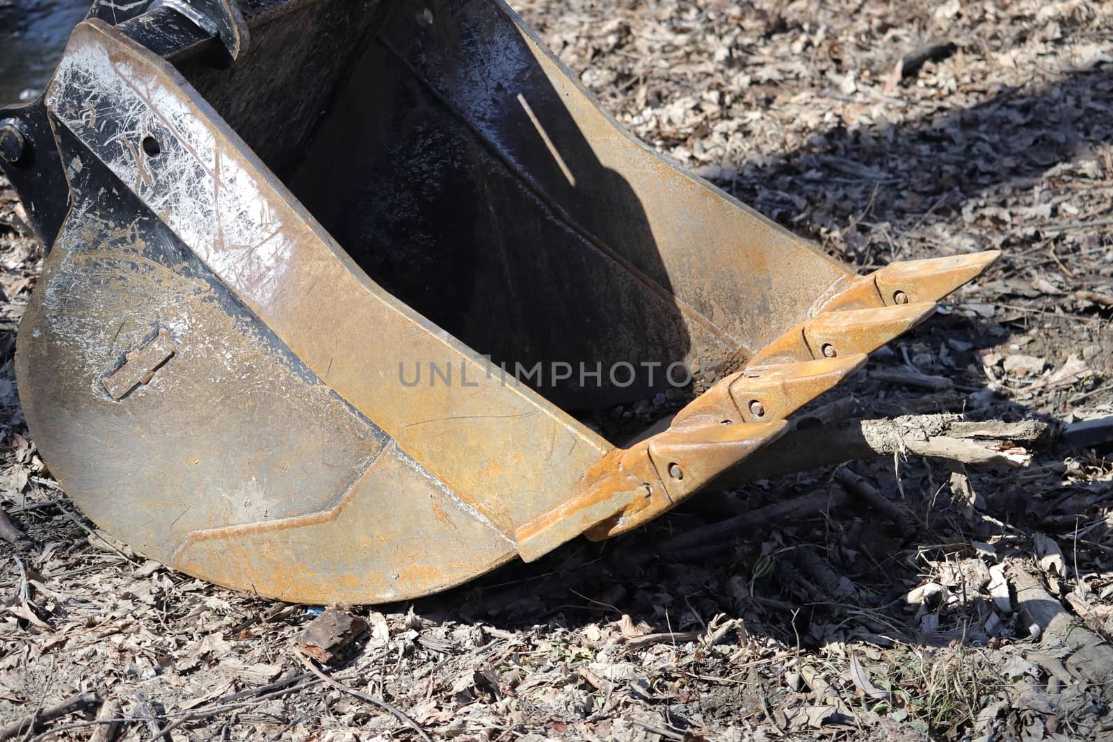 The rusty metal bucket, or shovel, of an excavator is detached from the machine and sitting on the ground, which is covered in dirty leaves and debris.