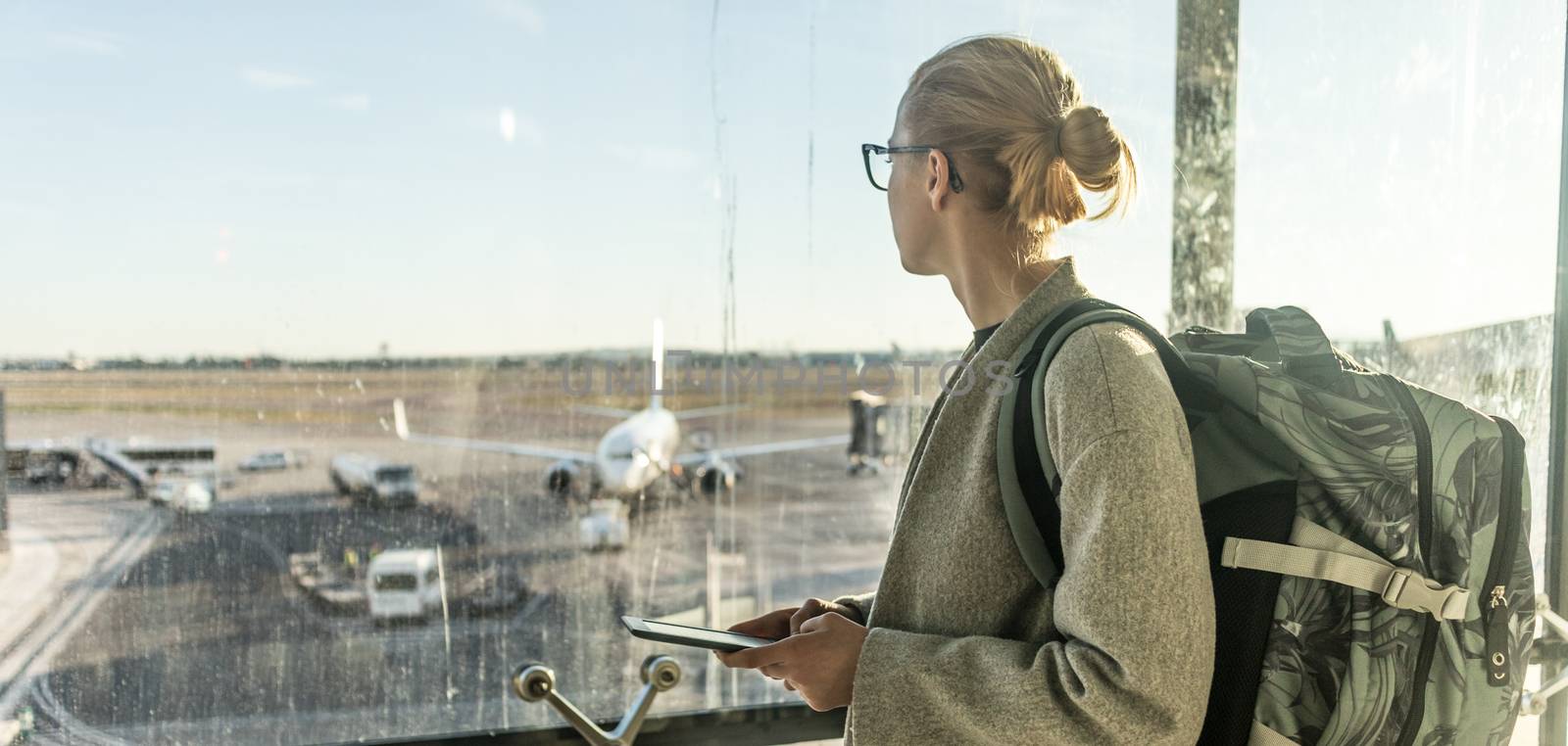 Young woman standing near airport gates window holding cellphone in her hands, wearing travel backpack and walking to lounge area. Female traveler search online map at the airport by kasto