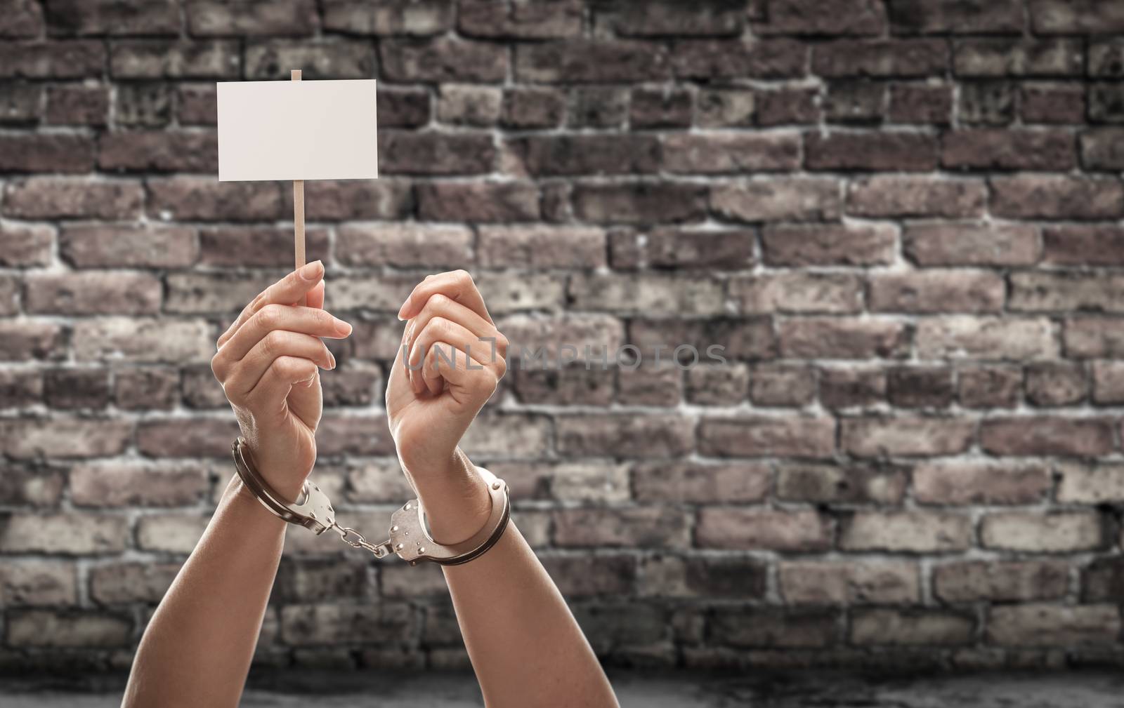 Handcuffed Female Hand Holding Blank Sign Against Aged Brick Wal by Feverpitched