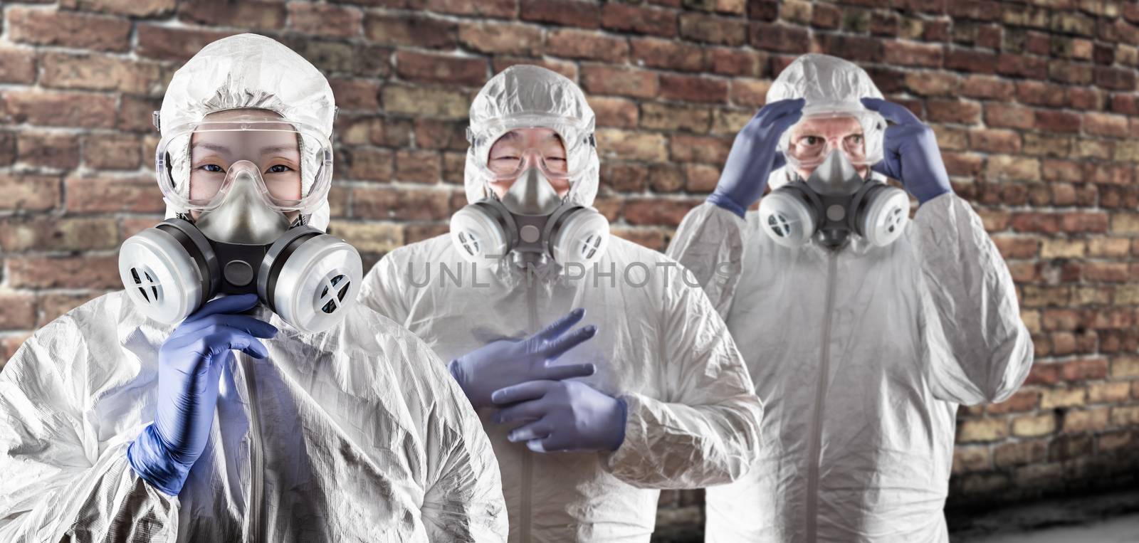 Chinese and Caucasian Woman and Men In Gas Masks, Goggles and Hazmat Suites Against Brick Background. by Feverpitched