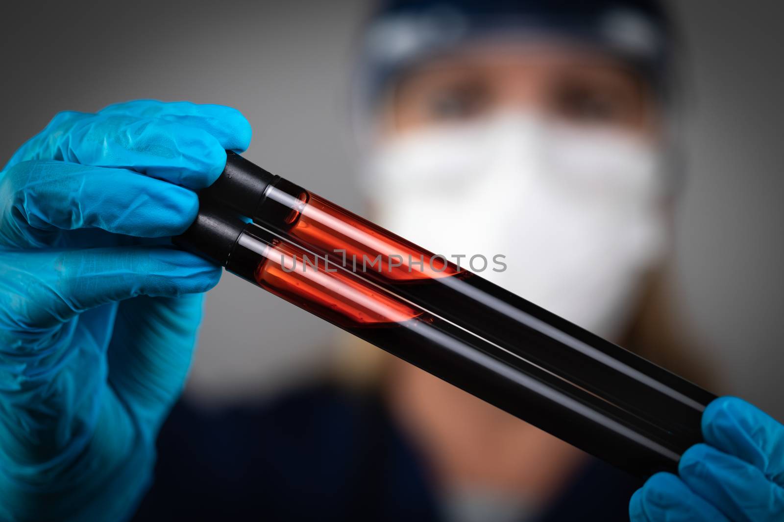 Female Lab Worker Wearing Medical Face Mask Holds Test Tubes of Blood Against Dark Background. by Feverpitched