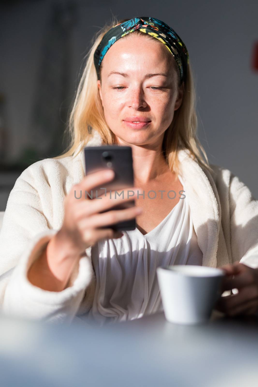 Beautiful caucasian woman at home, feeling comfortable wearing white bathrobe, taking some time to herself, drinking morning coffee and reading news on mobile phone device in the morning.