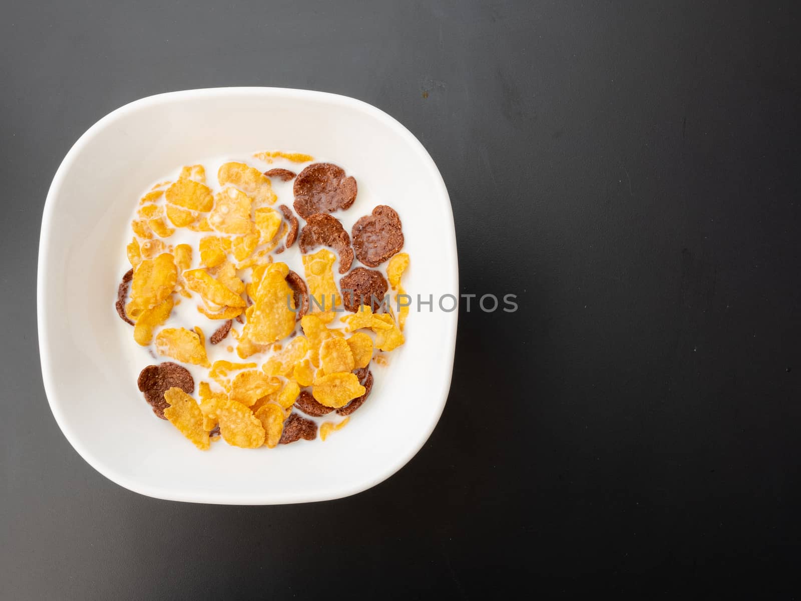 Breakfast is ready to be eaten in a white bowl consisting of cornflakes, crispy chocolate, and milk, placed on a black painted metal dining table.