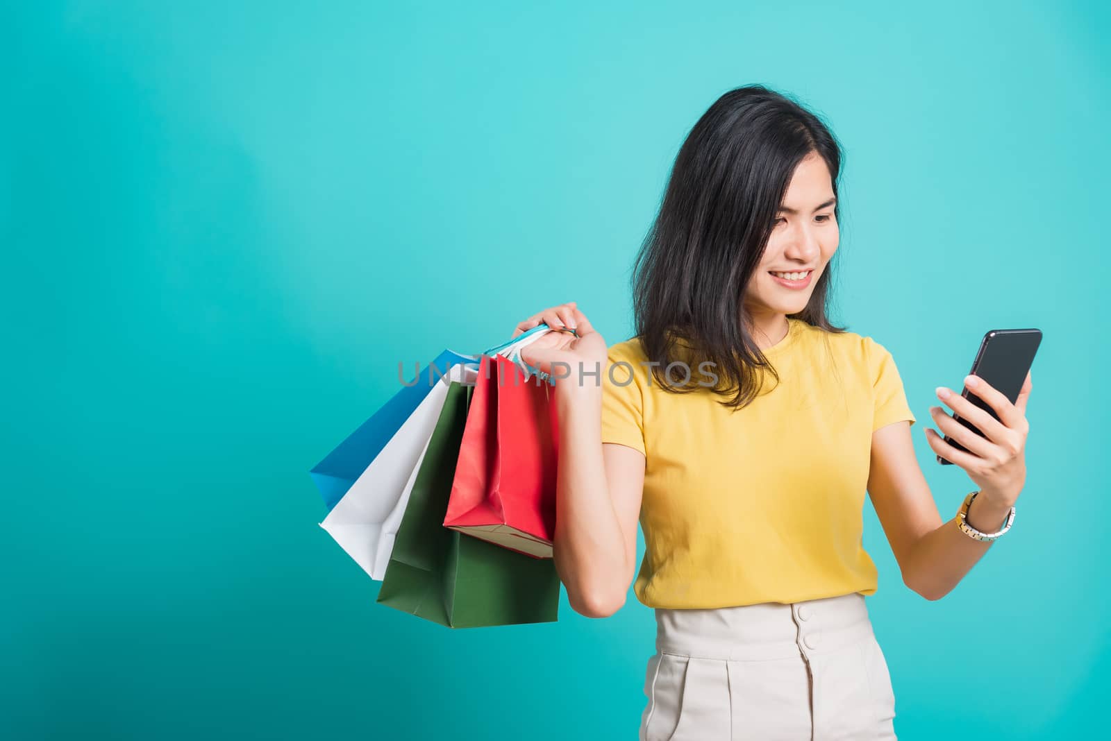 Portrait happy Asian beautiful young woman smile white teeth standing wear yellow t-shirt, She holding shopping bags and using a mobile phone, studio shot on blue background with copy space for text
