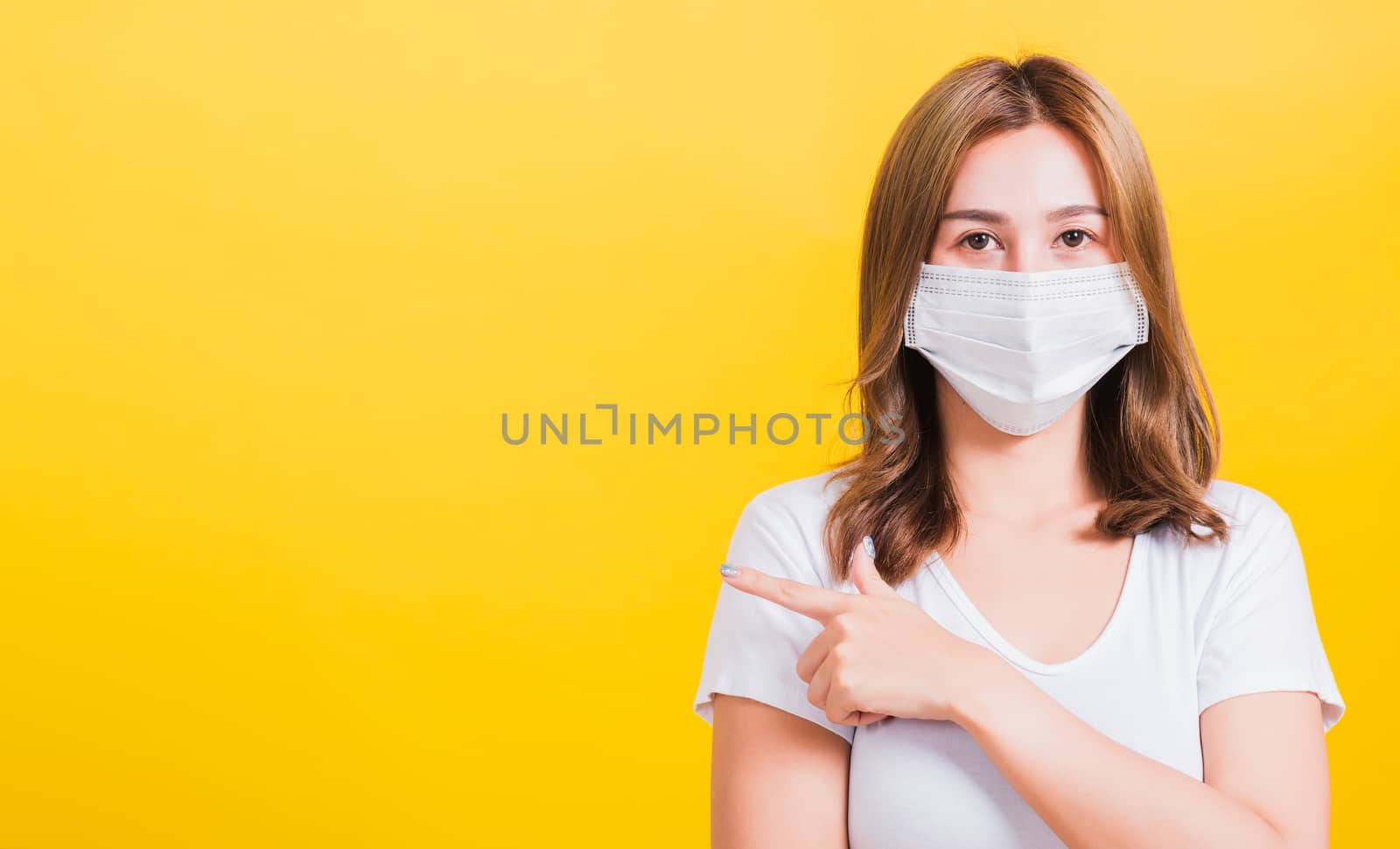 Portrait Asian beautiful young woman wearing face mask protects filter dust pm2.5 anti-pollution, anti-smog, air pollution and COVID virus her pointing out side on yellow background, with copy space