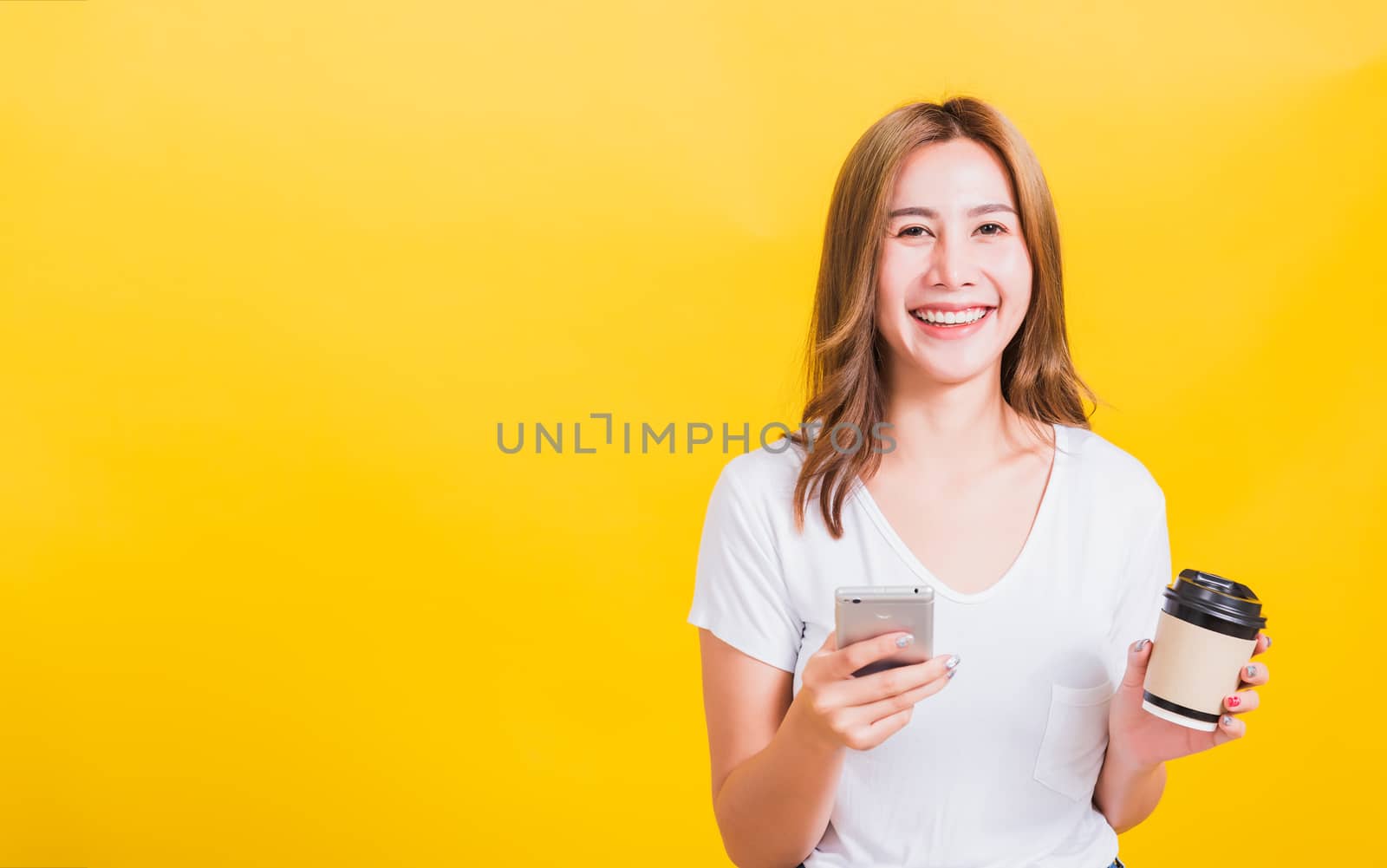 Portrait Asian Thai beautiful happy young woman standing smile, using mobile phone her holding coffee paper cup, looking to camera, studio shot isolated on yellow background, with copy space