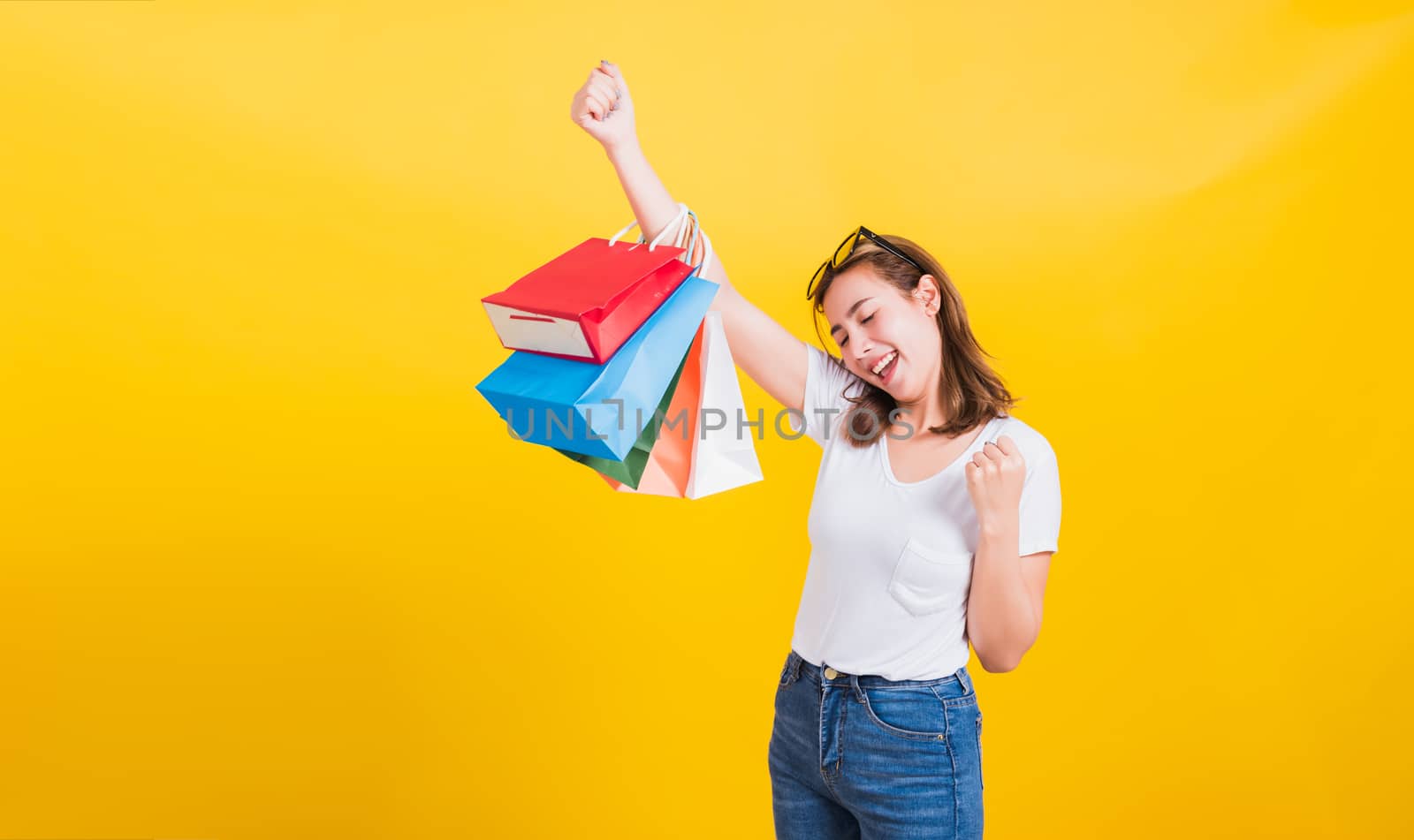 Asian Thai portrait happy beautiful cute young woman smiling stand with sunglasses excited holding shopping bags multi color looking camera, studio shot isolated yellow background with copy space