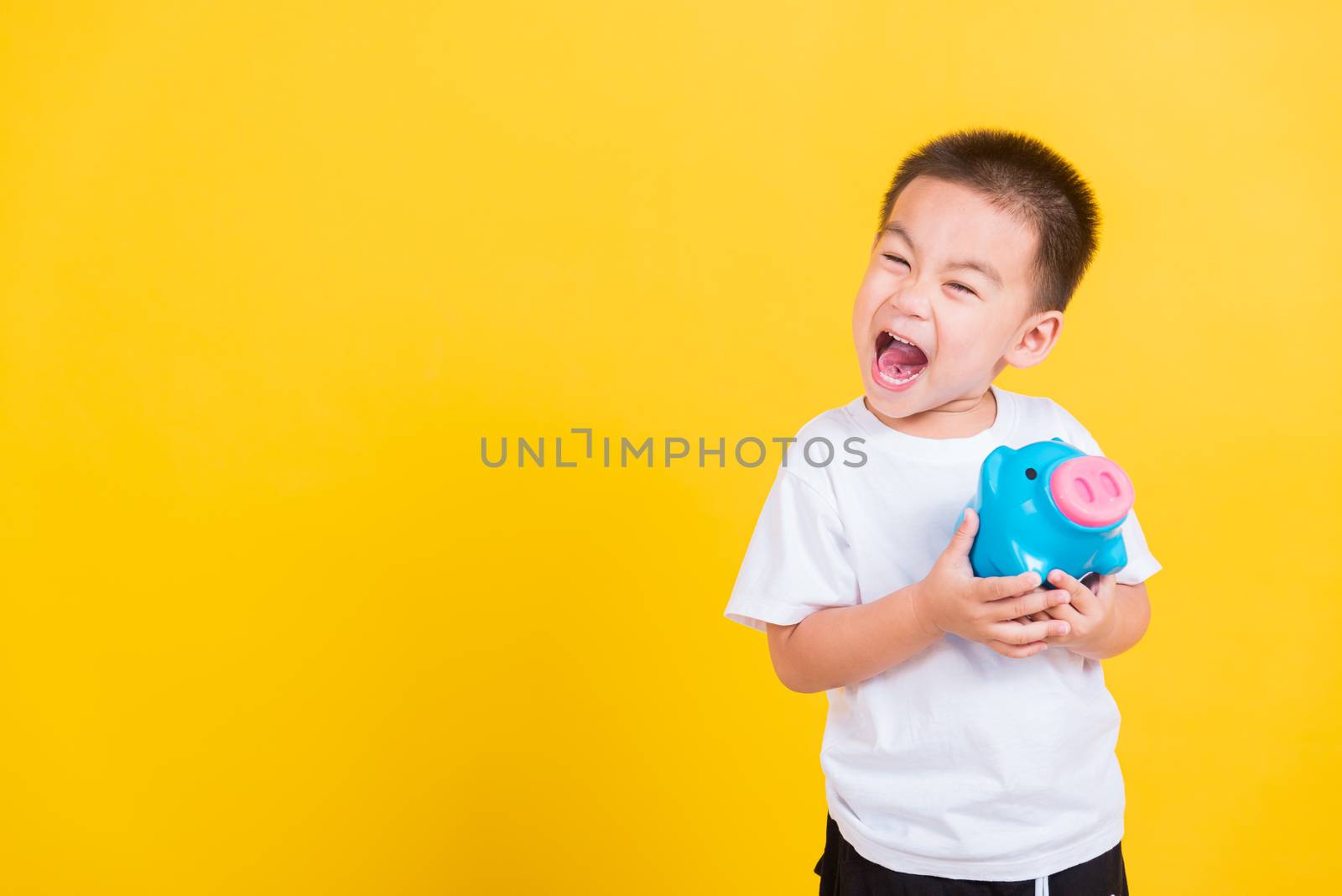 Asian Thai happy portrait cute little cheerful child boy smile holding piggy bank and looking camera, studio shot isolated on yellow background with copy space