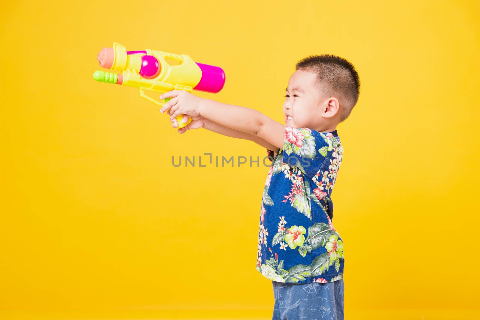 Portrait happy Asian cute little children boy smile standing so happy wearing flower shirt in Songkran festival day holding water gun, studio shot on yellow background with copy space