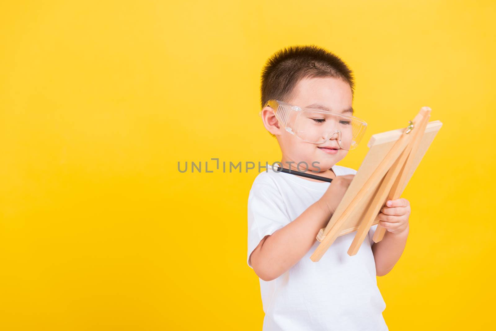 Asian Thai happy portrait cute little cheerful child boy smile are drawing on blackboard and looking the board, studio shot isolated on yellow background with copy space