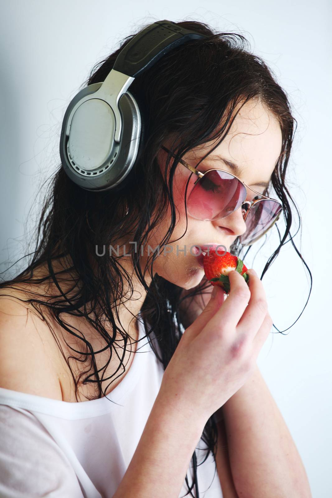Young woman listening to music on headphones eating strawberry. Closeup portrait of girl on light background with copy space