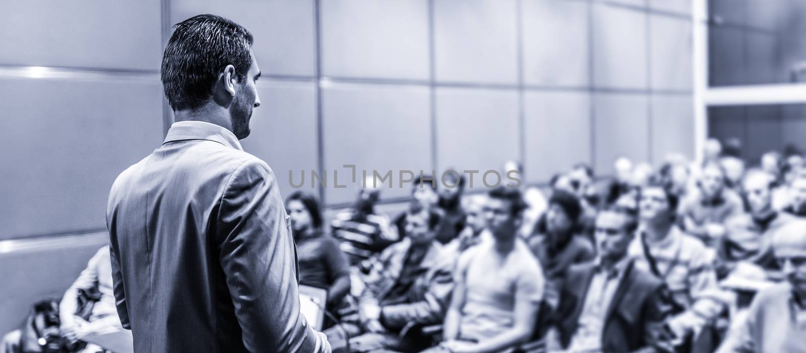 Speaker giving a talk on corporate business conference. Unrecognizable people in audience at conference hall. Business and Entrepreneurship event. Desaturated blue toned.