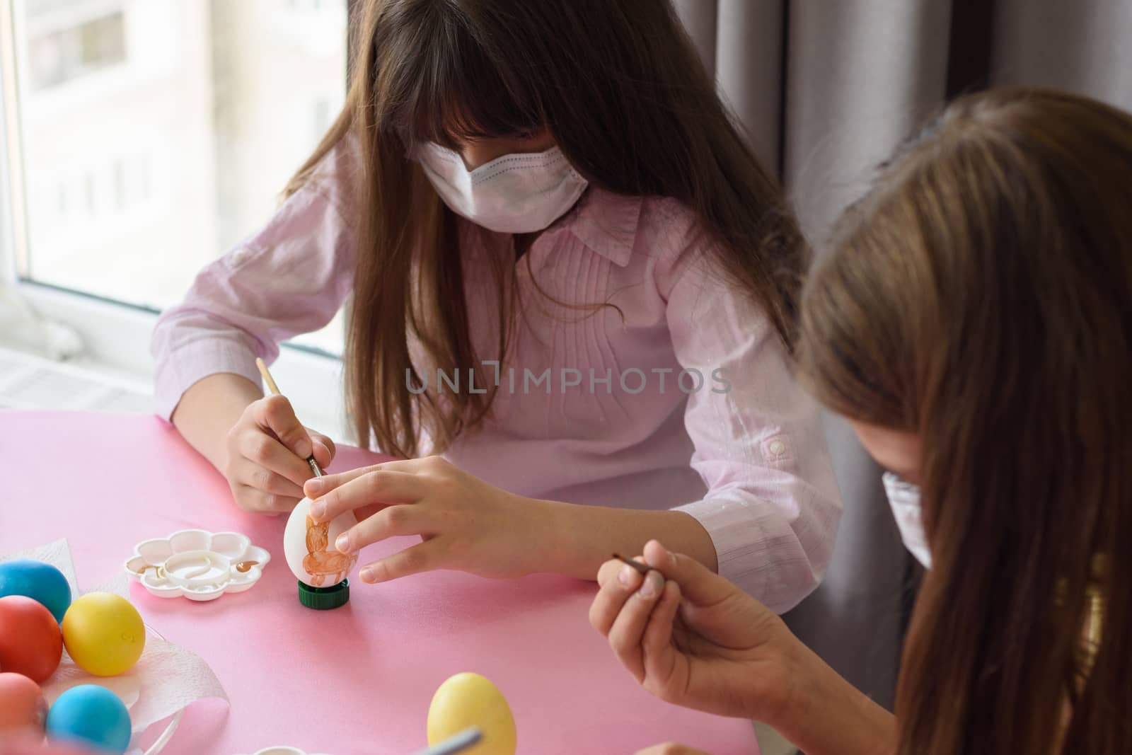 Two girls in medical masks paint eggs with a brush while preparing for Easter