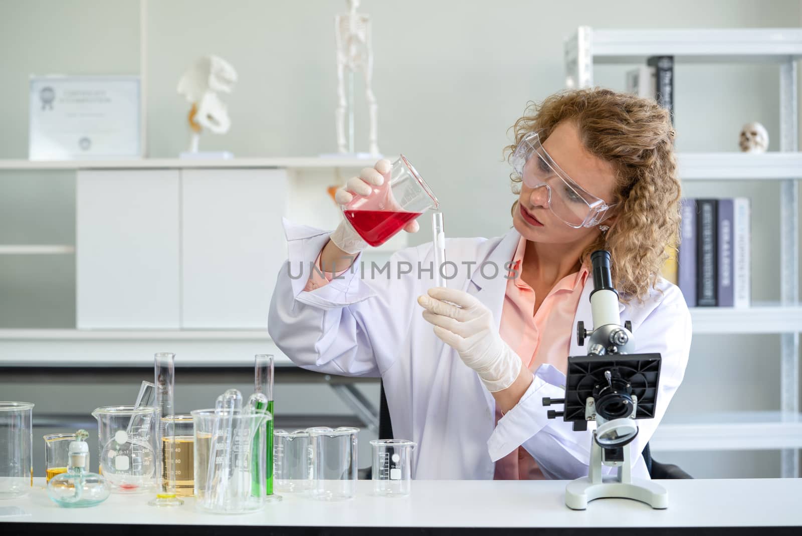 A young blonde scientist is pouring red chemicals into a test tube. Working atmosphere in chemical laboratory. Test tubes and beakers filled with chemicals on the table.