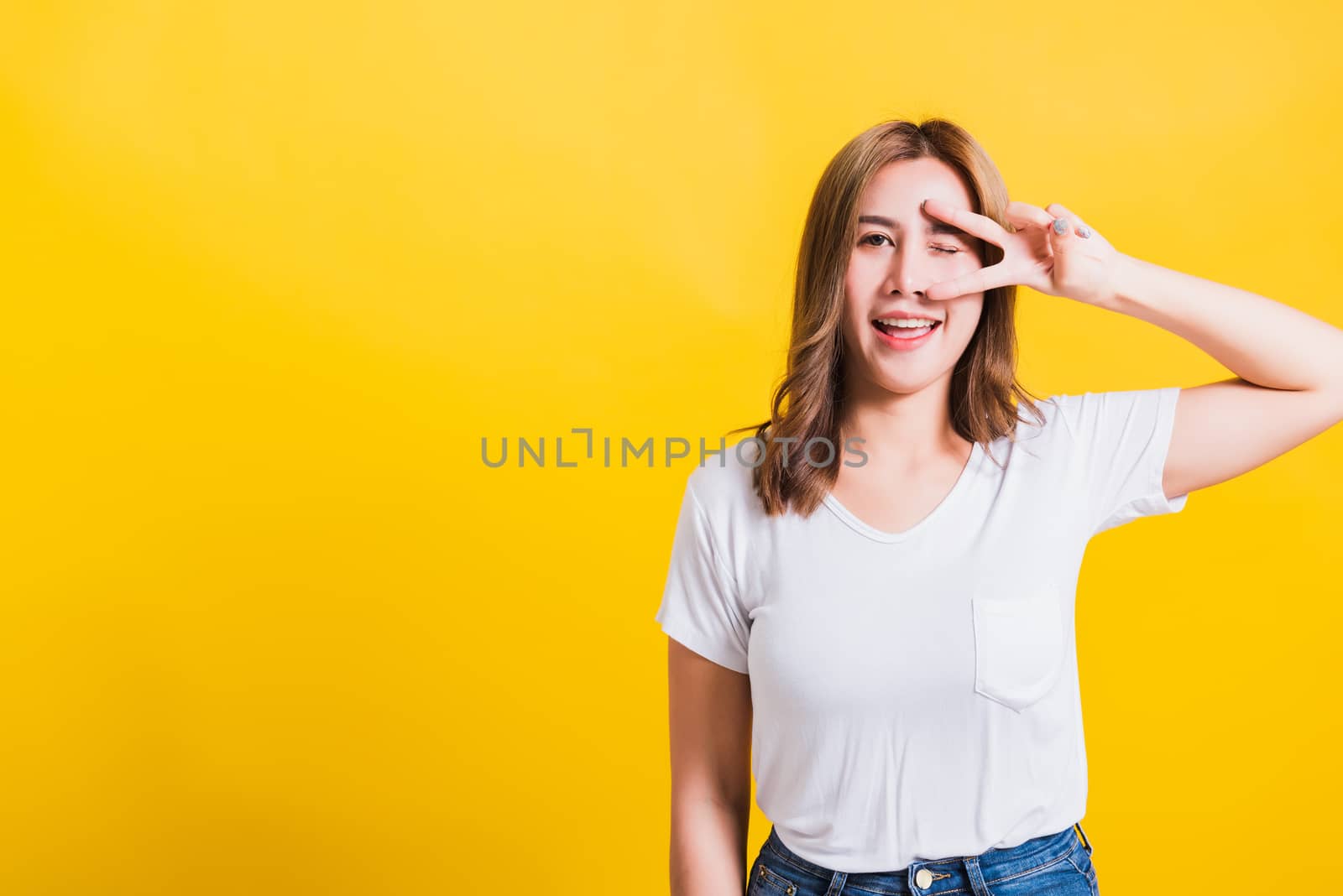 Asian Thai happy portrait beautiful cute young woman smile standing wear t-shirt showing finger making v-sign symbol near eye looking to camera, isolated studio shot yellow background with copy space