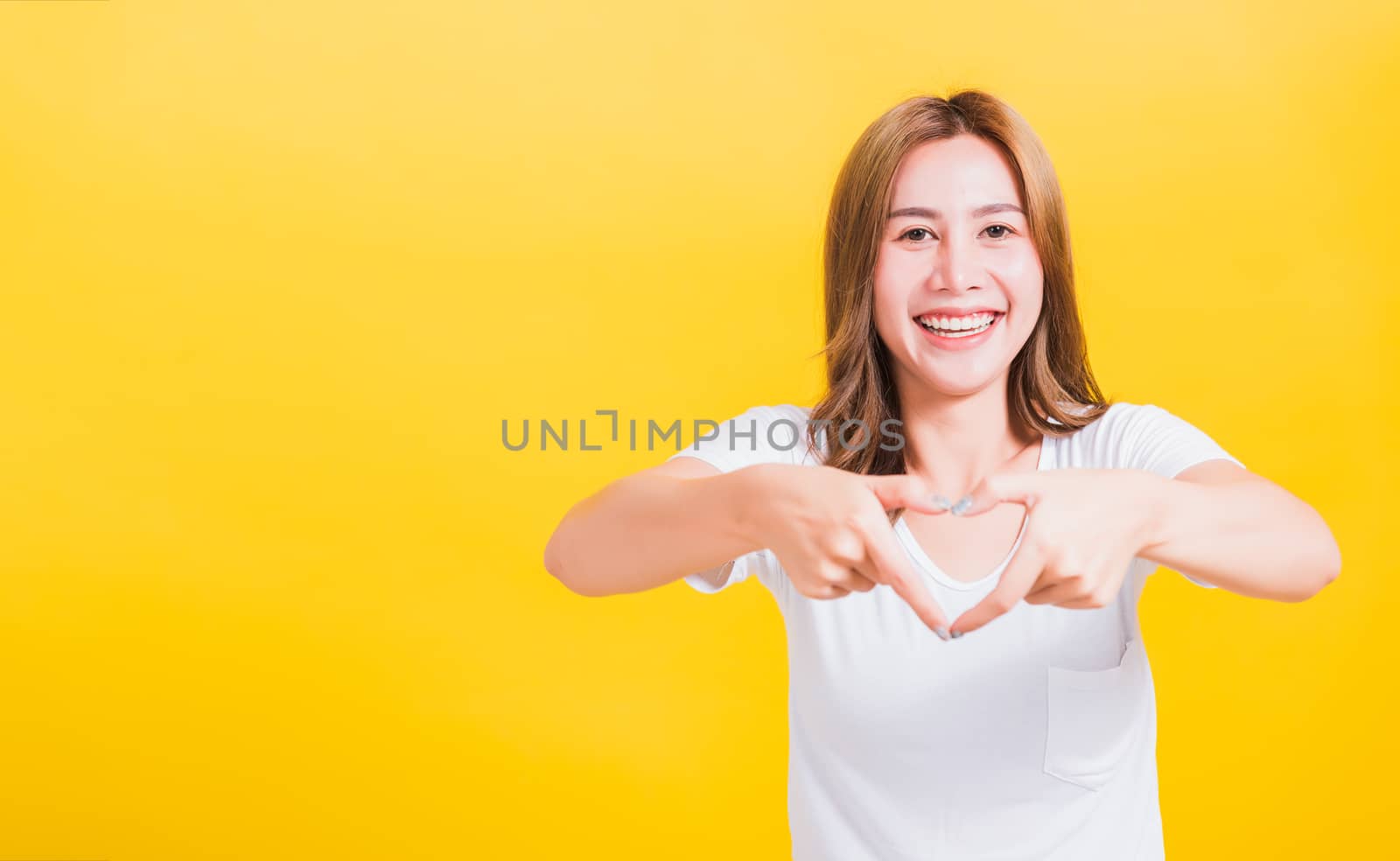 Asian Thai happy portrait beautiful cute young woman smile standing make finger heart figure symbol shape sign with two hands and looking camera, studio shot isolated yellow background with copy space