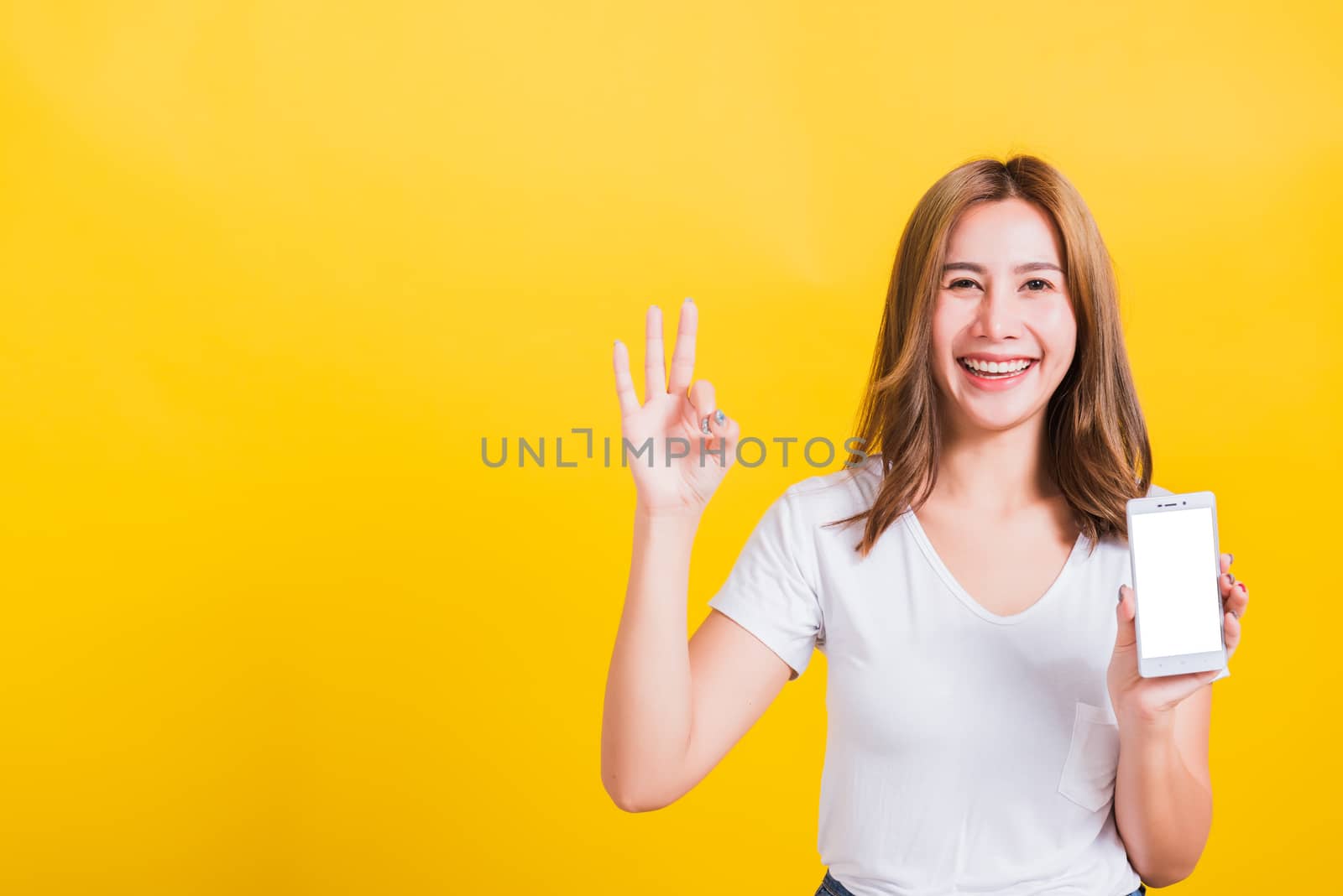 Portrait Thai Asian beautiful happy young woman stand smile, holding mobile phone blank screen and showing OK gesture, shoot photo in studio focus face isolated on yellow background, with copy space