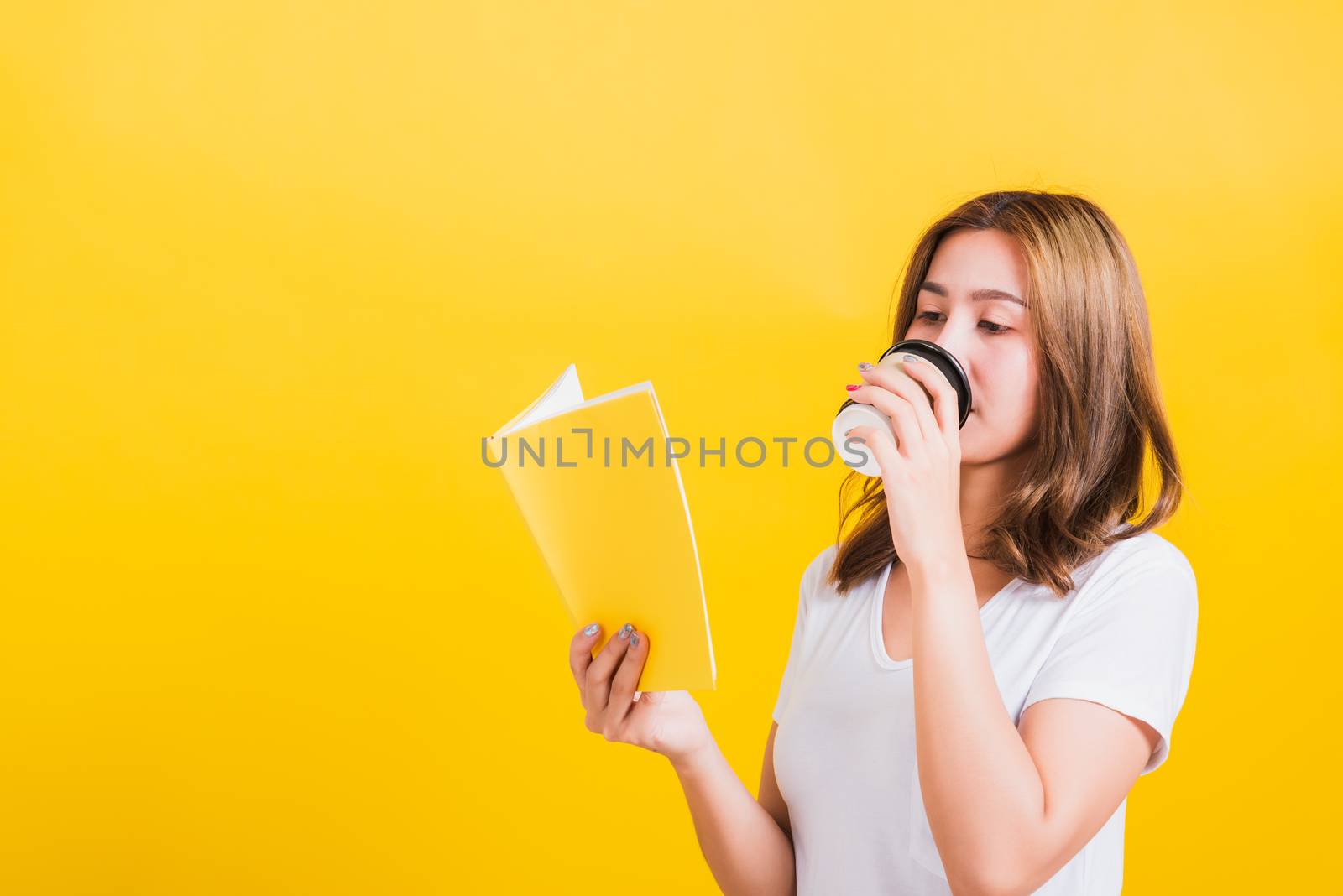 young woman standing holding drinking take away cup coffee paper and reading yellow book by Sorapop