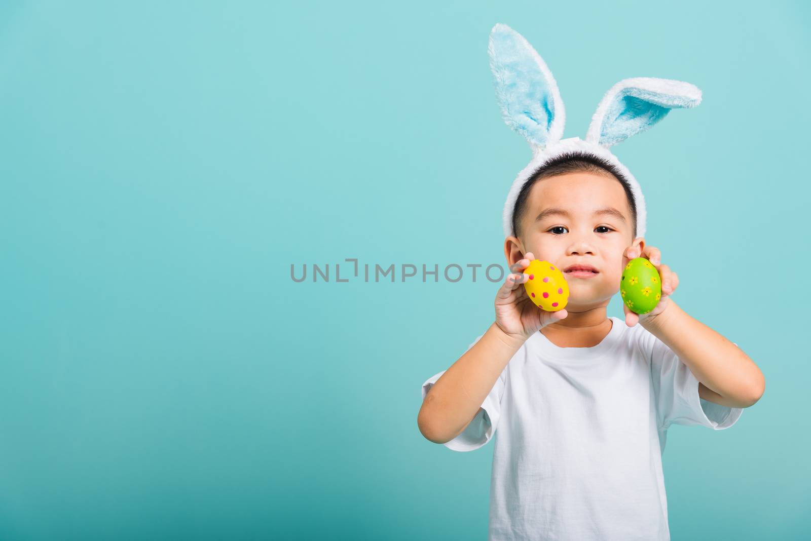 Asian cute little child boy smile beaming wearing bunny ears and a white T-shirt, standing using hand holding and show colored easter eggs on blue background with copy space