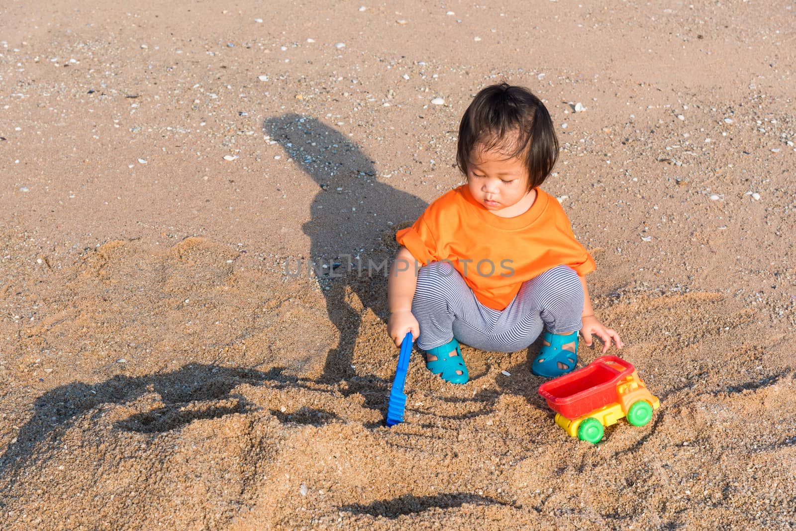 Asian Thai happy cute little cheerful daughter girl funny digging playing toy with sand at an outdoor tropical beach in summer day with copy space