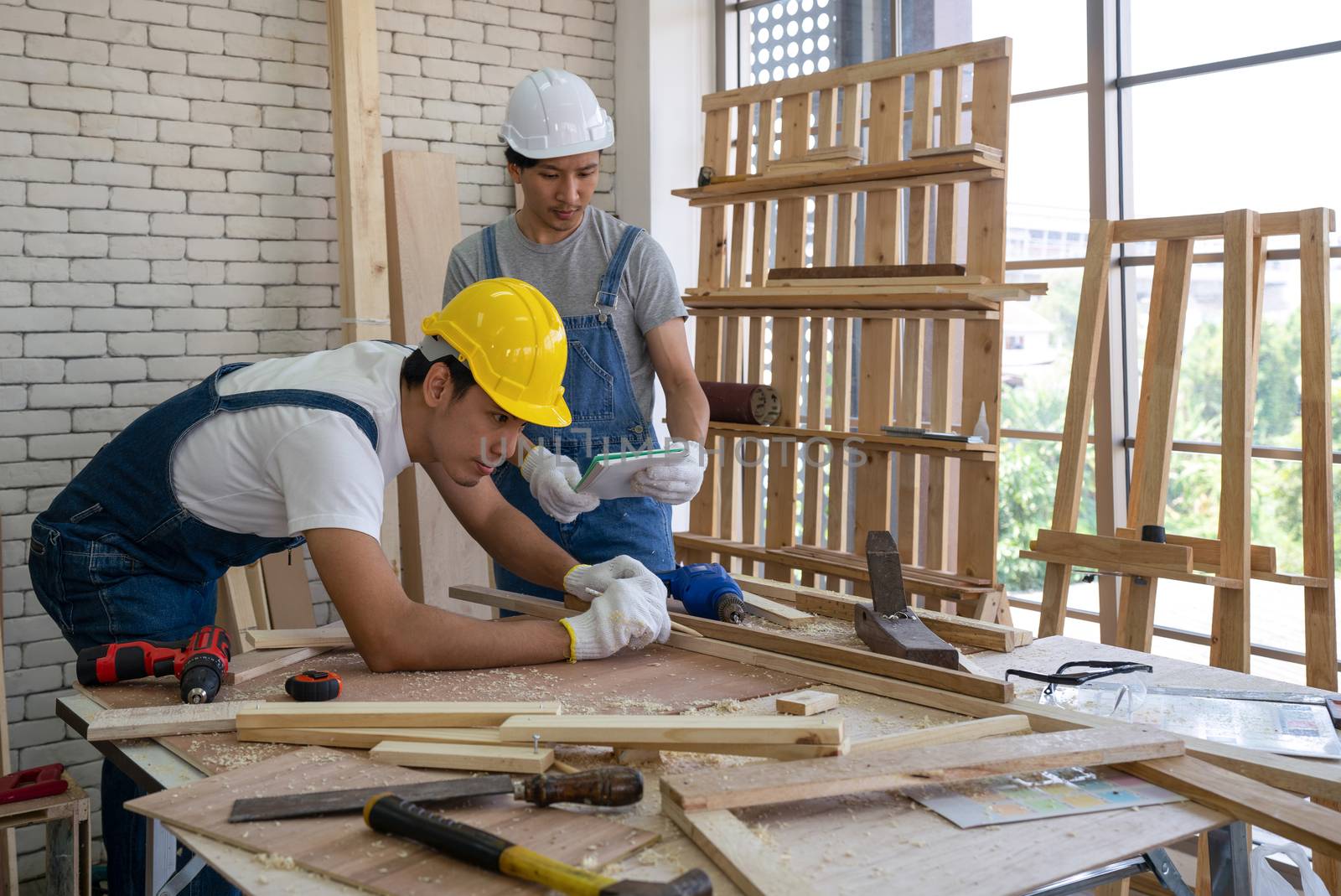 An Asian carpenter is drawing a line on a piece of wood, preparing to cut into sections. The other man told the size he wanted by looking at his note. Morning work atmosphere in the workshop room.