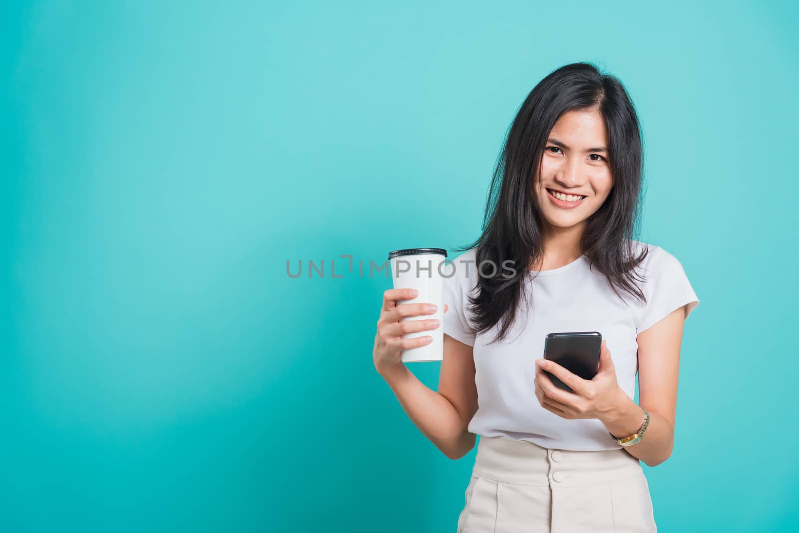 Portrait Asian beautiful happy young woman standing smile, using mobile phone her holding coffee paper cup, looking to camera on blue background, with copyspace