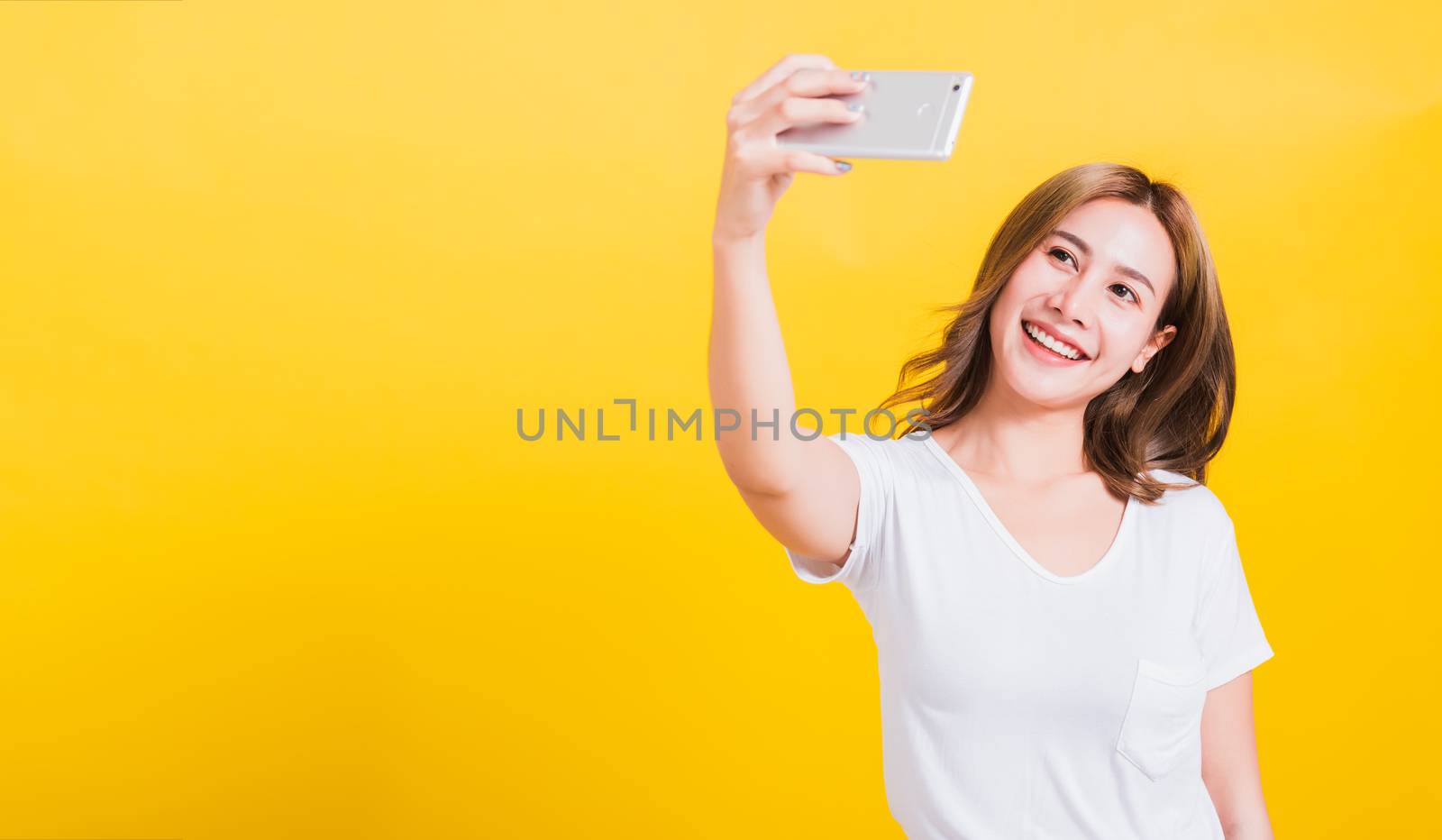 Asian Thai portrait happy beautiful cute young woman smiling wear white t-shirt making selfie photo, video call on smartphone looking the phone, studio shot isolated yellow background with copy space