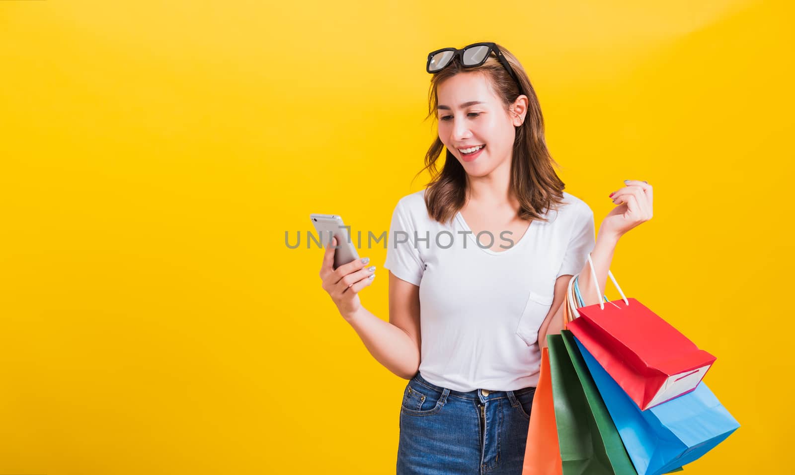 Portrait happy Asian Thai beautiful young woman smile white teeth stand wear t-shirt, She holding shopping bags and using app in mobile phone, studio shot isolated on yellow background with copy space