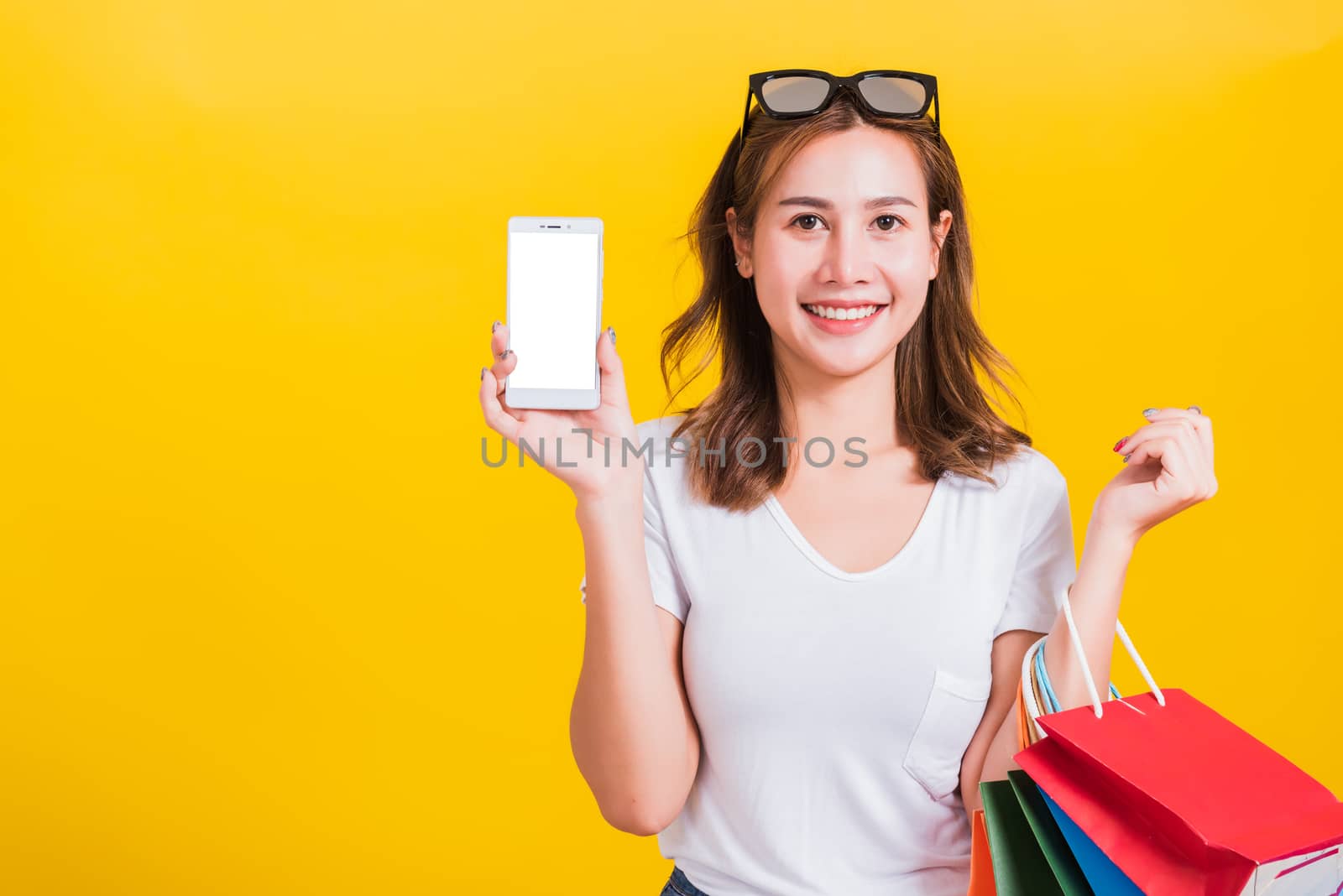 Portrait happy Thai Asian beautiful young woman smile white teeth standing wear t-shirt, She holding shopping bags and show mobile phone blank screen, studio shot on yellow background with copy space