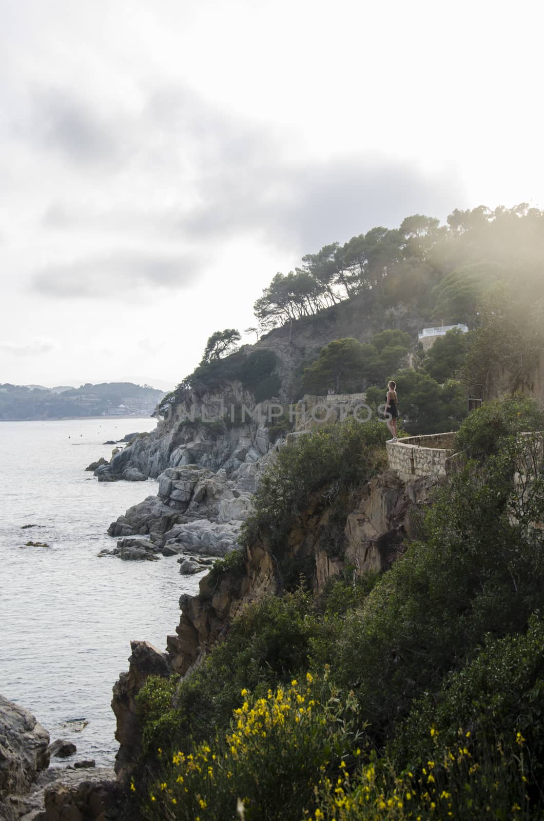 Attractive young woman in black dress and with a yellow little bag looking on a sea in sunset. Slim beautiful girl dress stands against rolling sea and beautiful rocks in Spain, Lloret de Mar