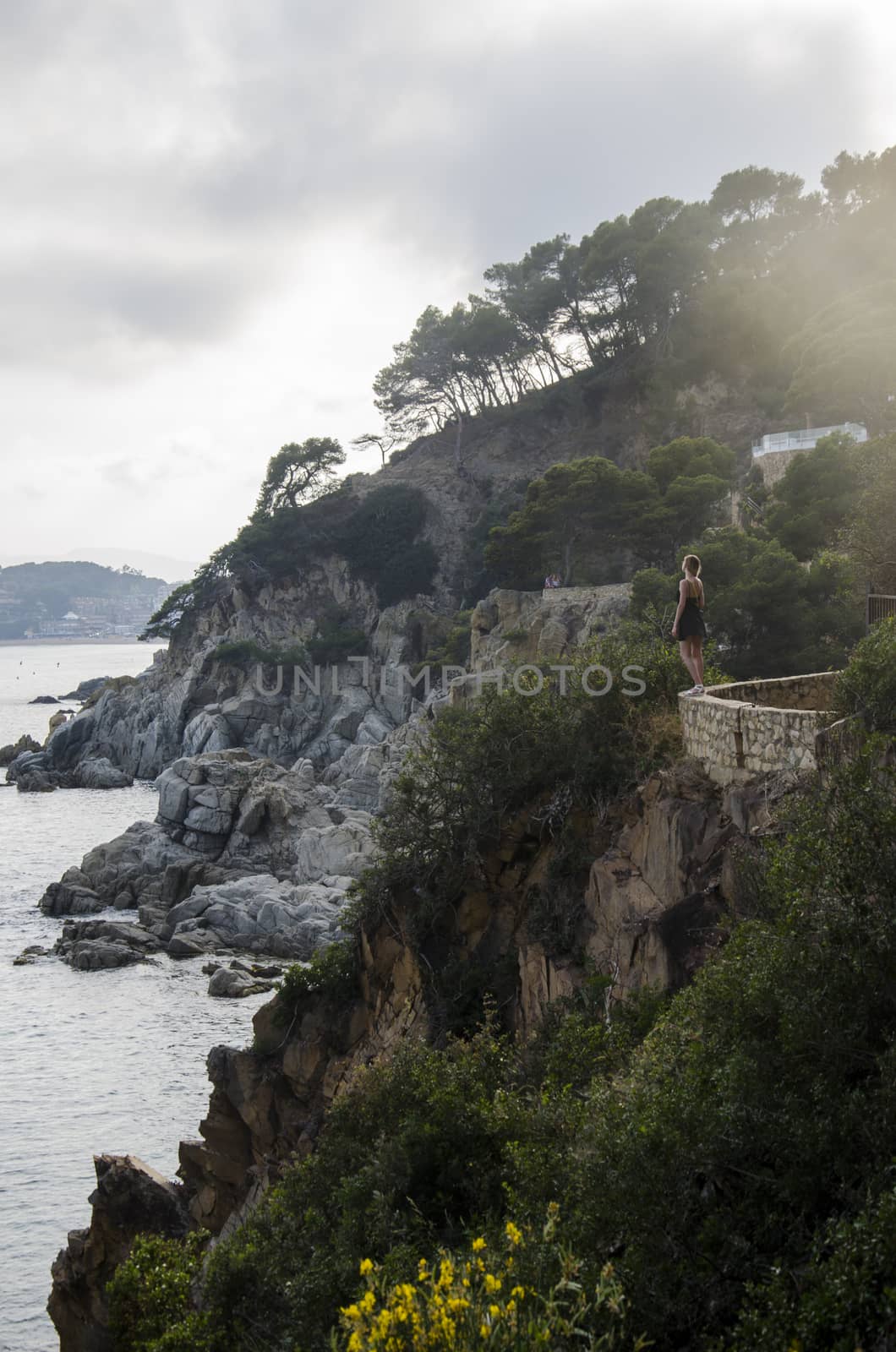 Attractive young woman in black dress and with a yellow little bag looking on a sea in sunset. Slim beautiful girl dress stands against rolling sea and beautiful rocks in Spain, Lloret de Mar. by vovsht