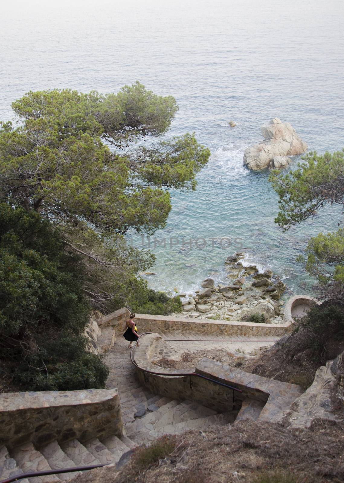 Panorama of Beautiful rocks stairs in Lloret de Mar and attractive young woman walking downstairs in black dress looking on a sea. Girl walking downstairs against rolling sea and rocks in Spain