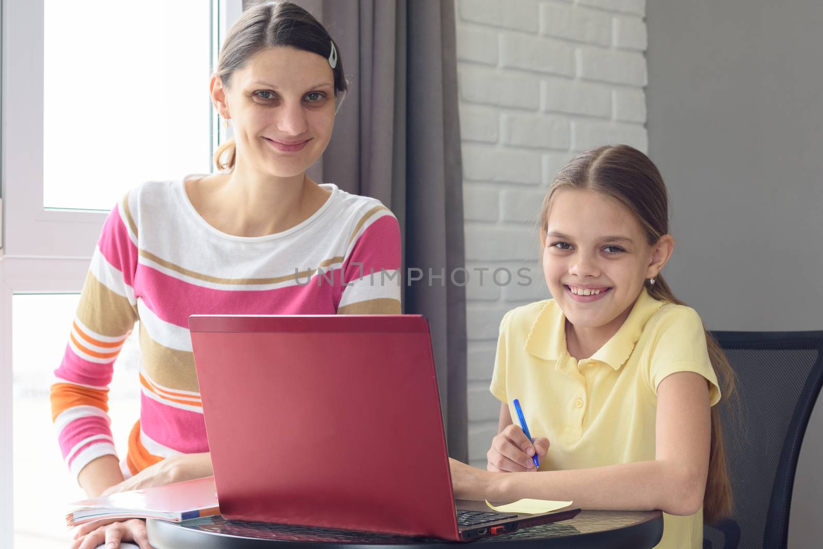 Happy girl and mom with a smile look at the frame, sitting at the table and doing homework