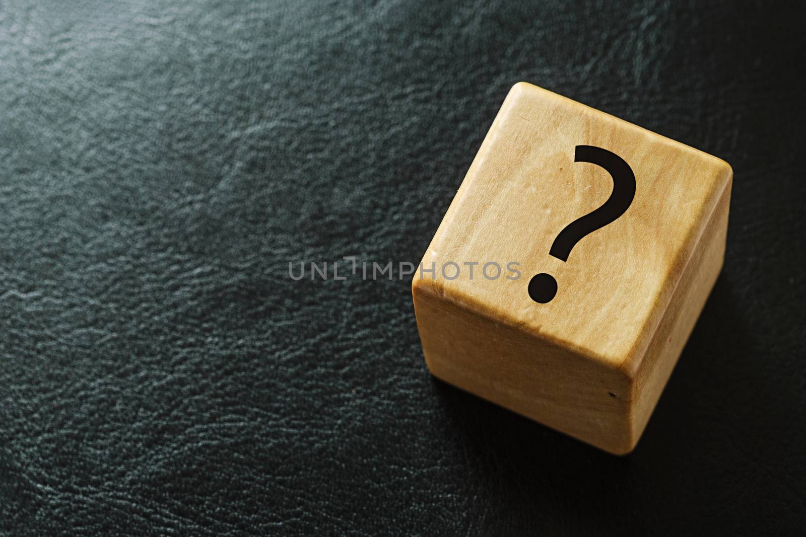 Wooden cube on black leather with question mark printed on one side viewed from above with copy space