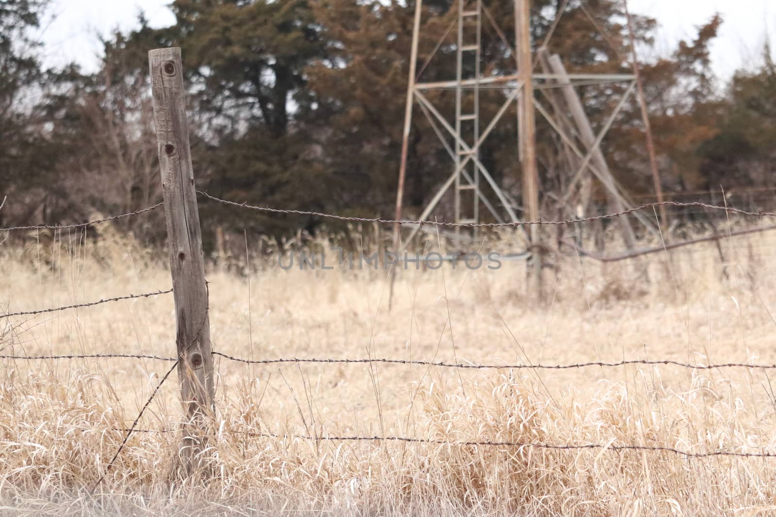 A close of a old wooden fence post and barbwire in a dry grass field with an old windmill