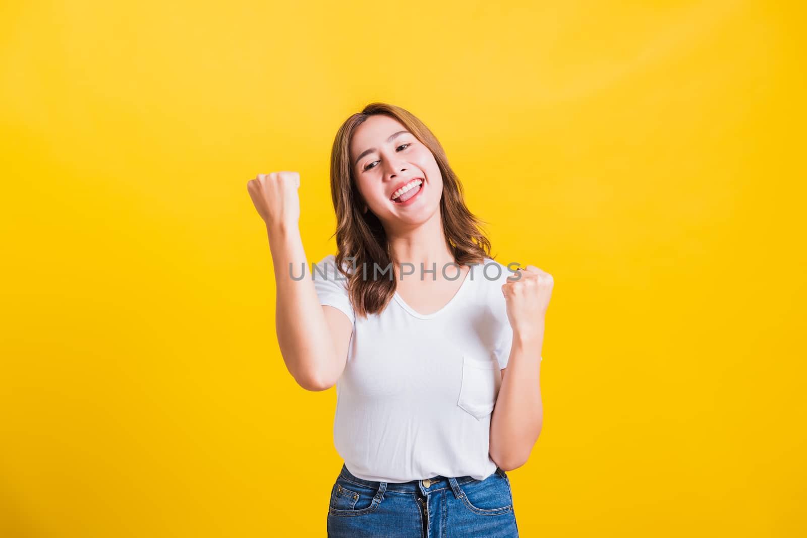 Asian Thai happy portrait beautiful cute young woman standing wear t-shirt makes raised fists up celebrating her winning success looking to camera isolated, studio shot on yellow background with copy space