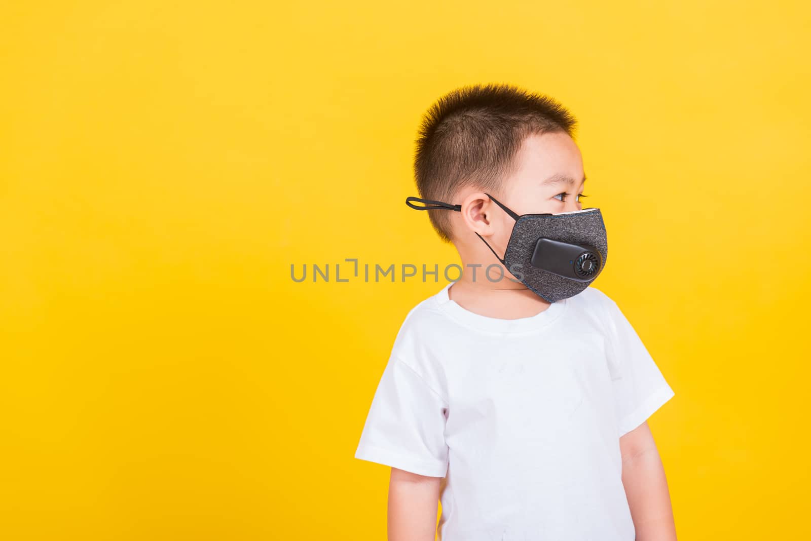 Asian Thai happy portrait cute little cheerful child boy wearing mask protective for covid-19 or pm2.5 dust he looking to side space, studio shot isolated on yellow background with copy space