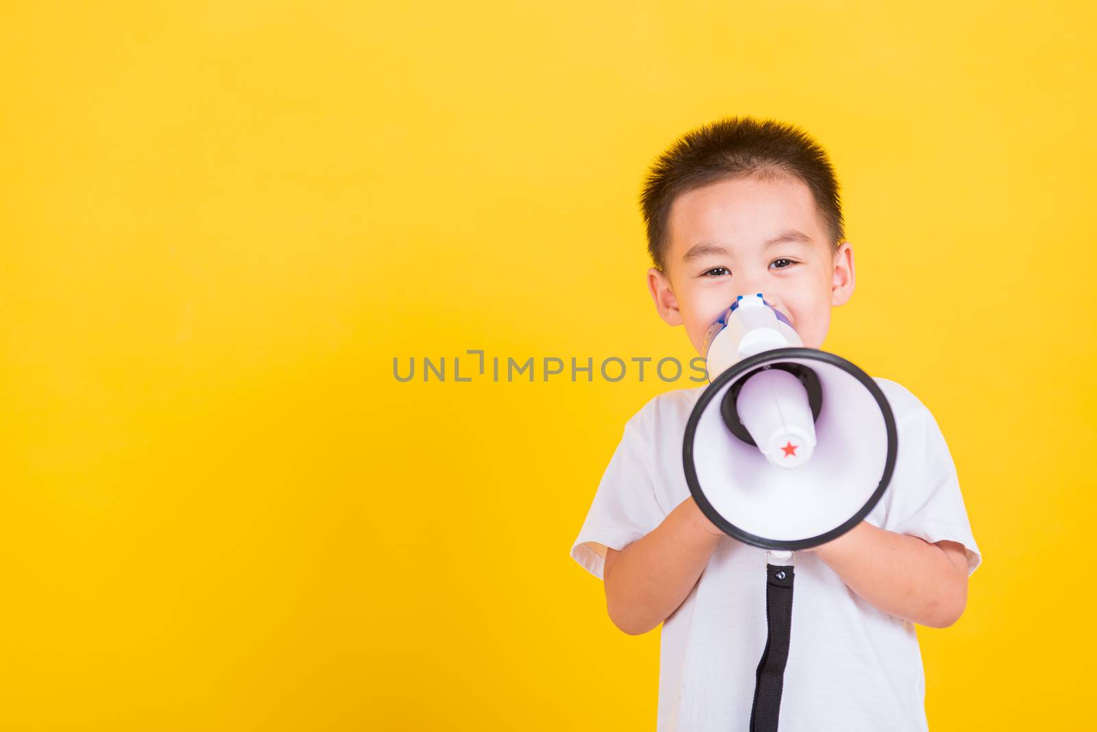 Asian Thai happy portrait cute little cheerful child boy holding and shouting or screaming through the megaphone her looking to camera, studio shot isolated on yellow background with copy space