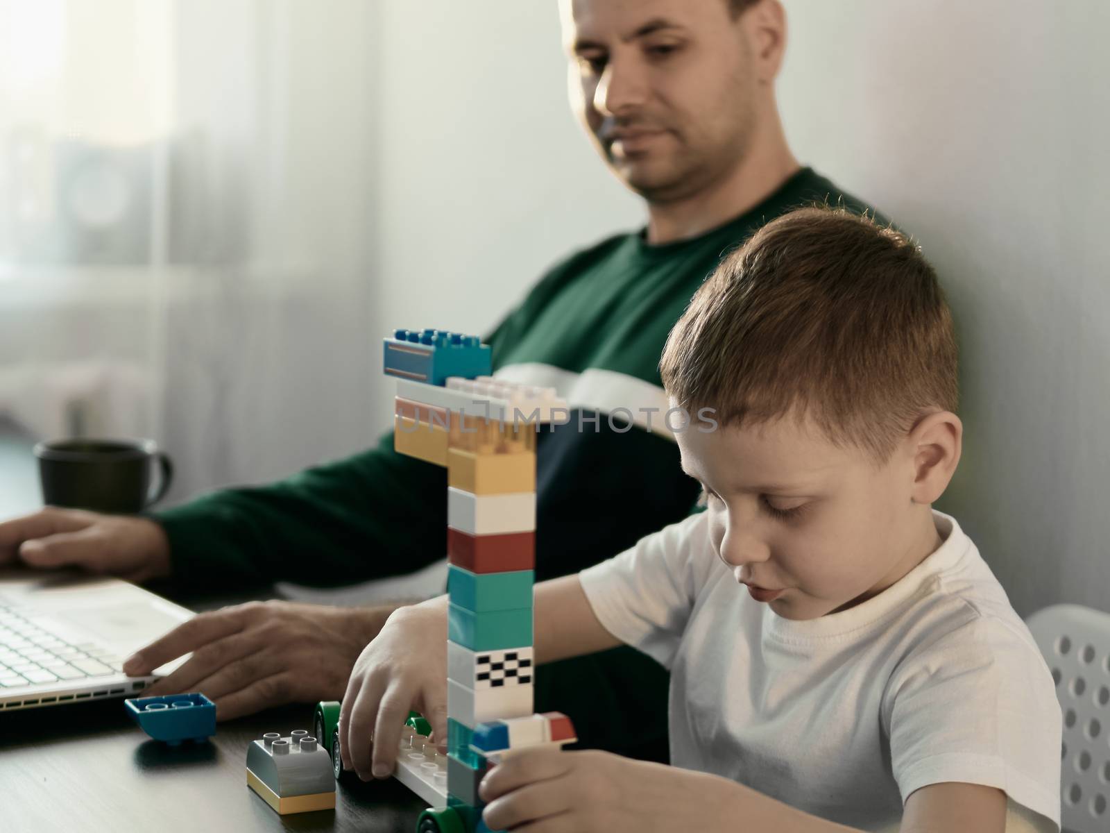 Four year old boy play with building set for children and dad who works at aptop at home. Home working at coronavirus quarantine isolation period. Working among children, working remote concept