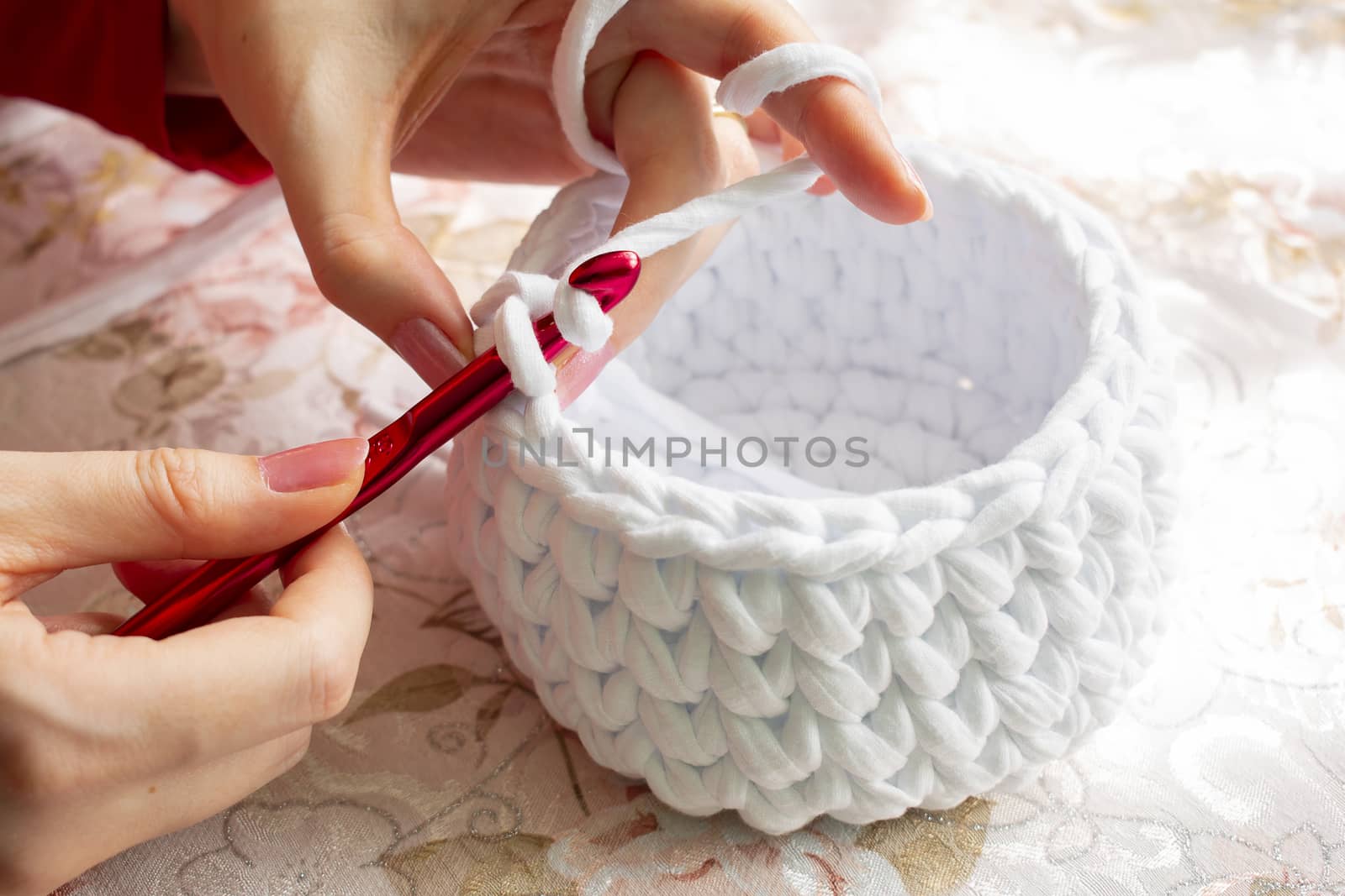 Young woman while crocheting on a patterned tablecloth, close up. Stay at home leisure activity idea. Basket made of white T-shirt yarn, with red crochet needle. Pale pink nails.
