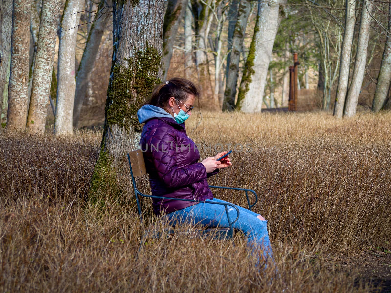 Lady and coronavirus protection. Sad Woman sitting alone on bench in park wearing mask to avoid infectious. Corona virus, or Covid 19, is spreading all over the world. Receives bad news on her phone.