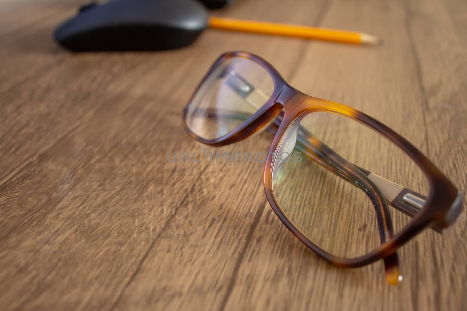 Closeup on a par of glasses with a pencil and mouse on the background, on top of a wooden table