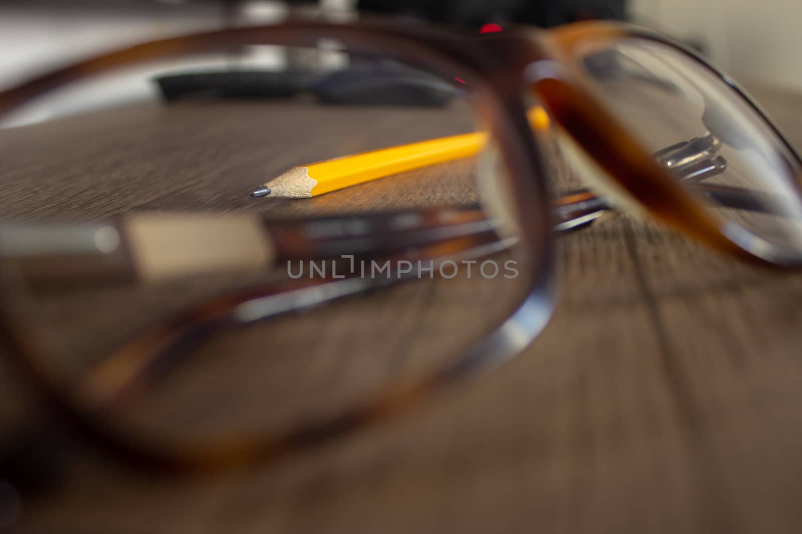 Focus on a yellow pencil through the lens of a par of glasses on top of a wooden desk, with a computer on the background