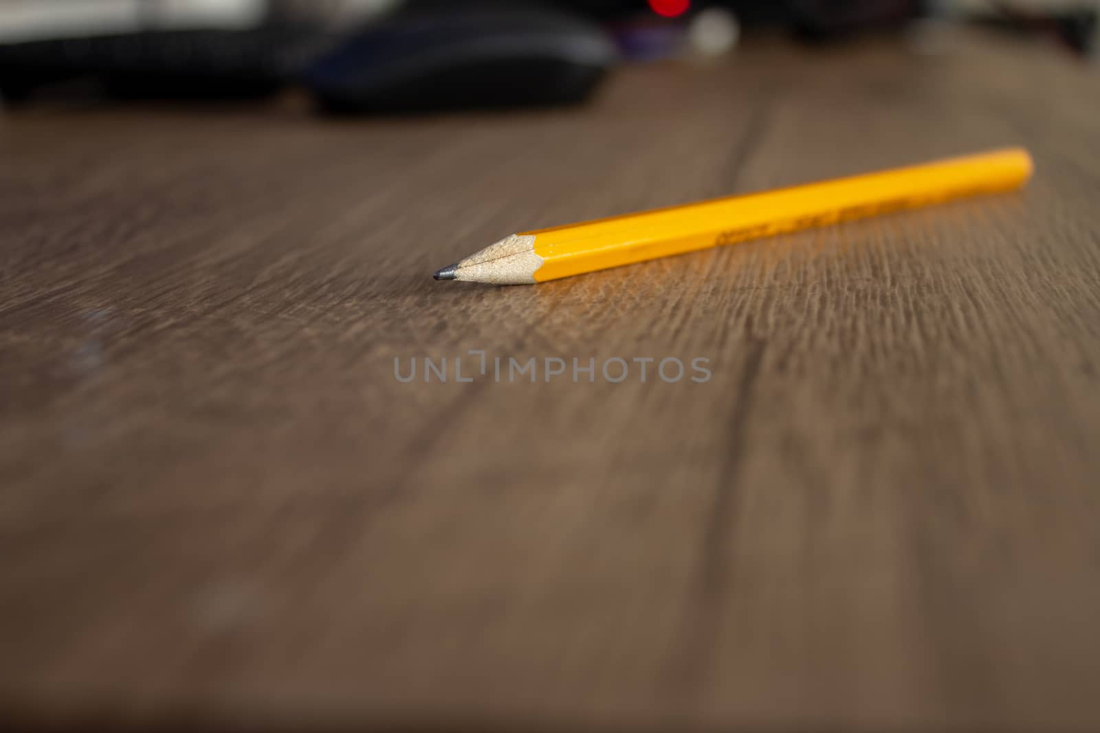 Focus on a yellow pencil on top of a wooden desk, with a computer on the background