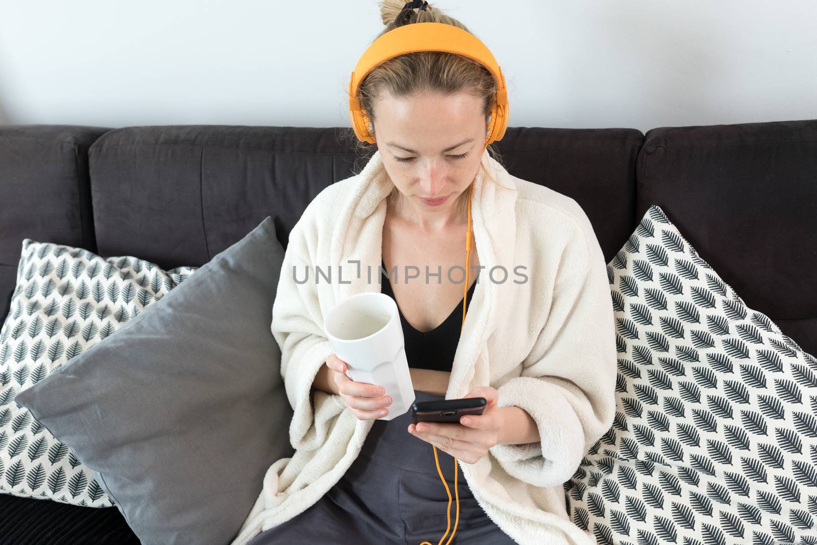 Stay at home. Social distancing. Woman at home relaxing on sofa couch drinking tea from white cup, listening to relaxing music, stay connected to friens and family via social networks on mobile. by kasto