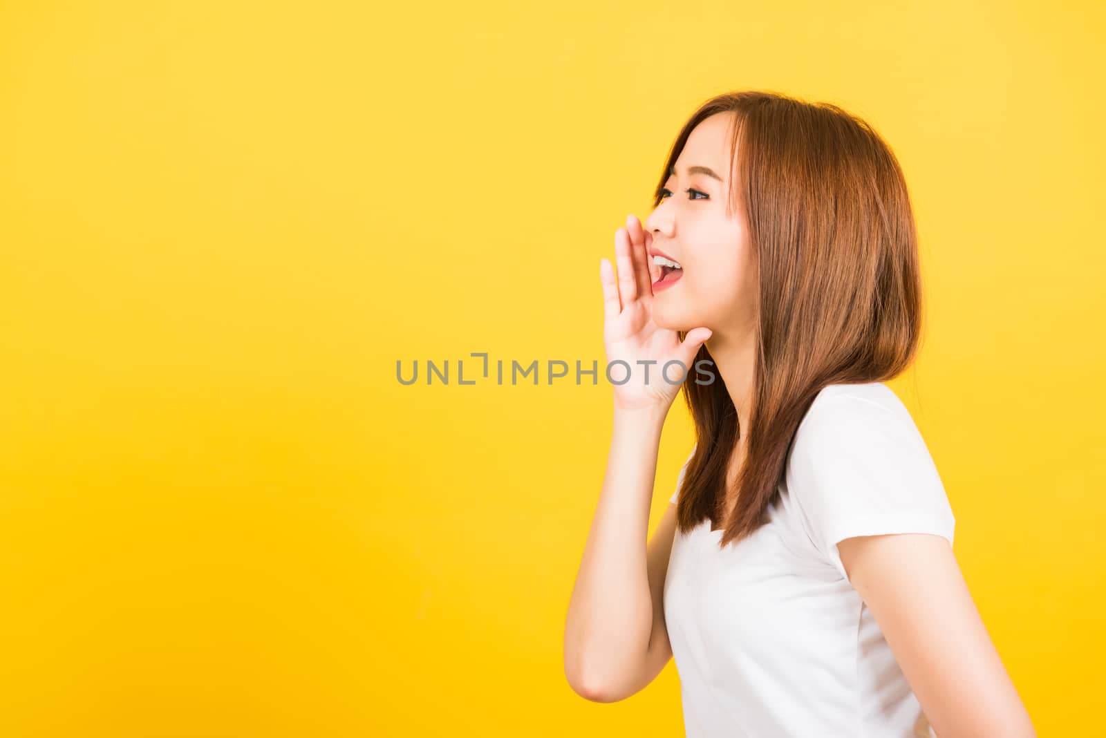 Asian happy portrait beautiful cute young woman teen standing wear t-shirt hand on mouth talking whispering secret rumor looking to side isolated, studio shot on yellow background with copy space