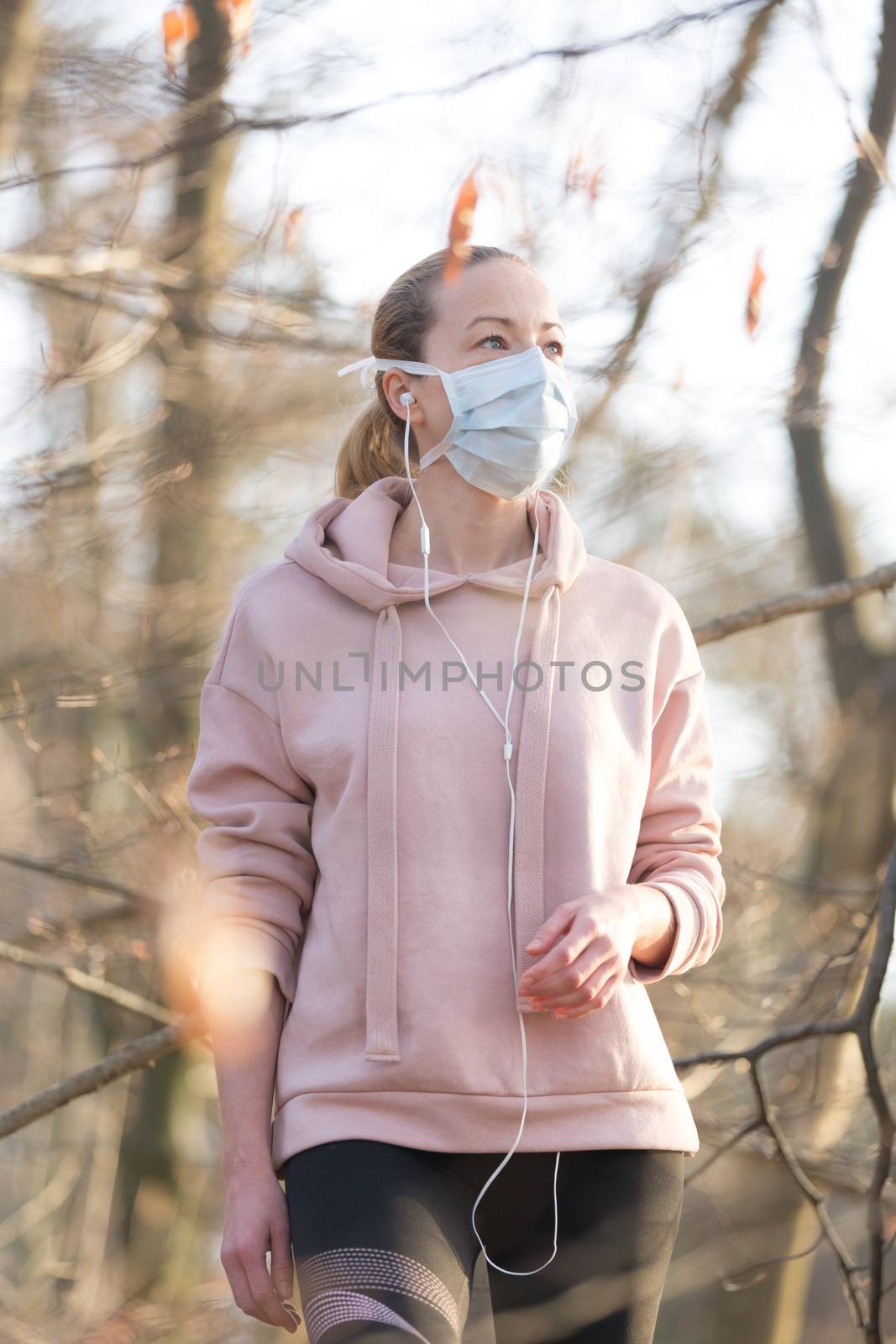 Portrait of caucasian sporty woman wearing medical protection face mask while walking in park, relaxing and listening to music. Corona virus, or Covid-19, is spreading all over the world.