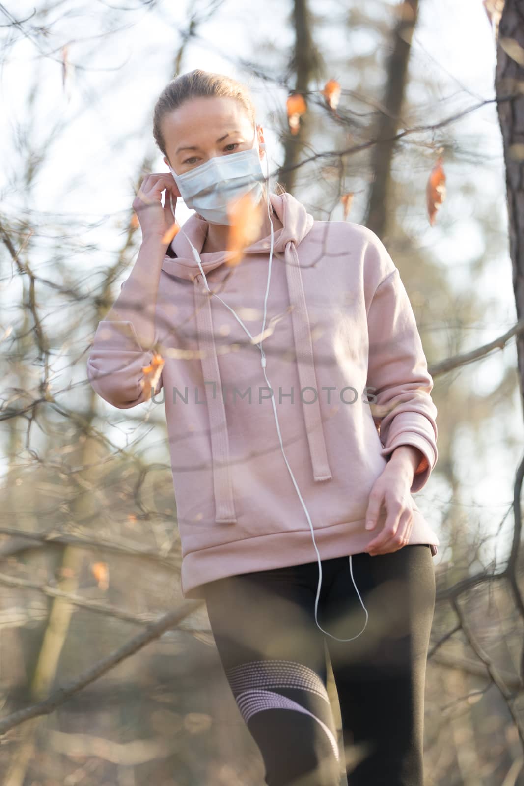 Portrait of caucasian sporty woman wearing medical protection face mask while walking in park, relaxing and listening to music. Corona virus, or Covid-19, is spreading all over the world by kasto