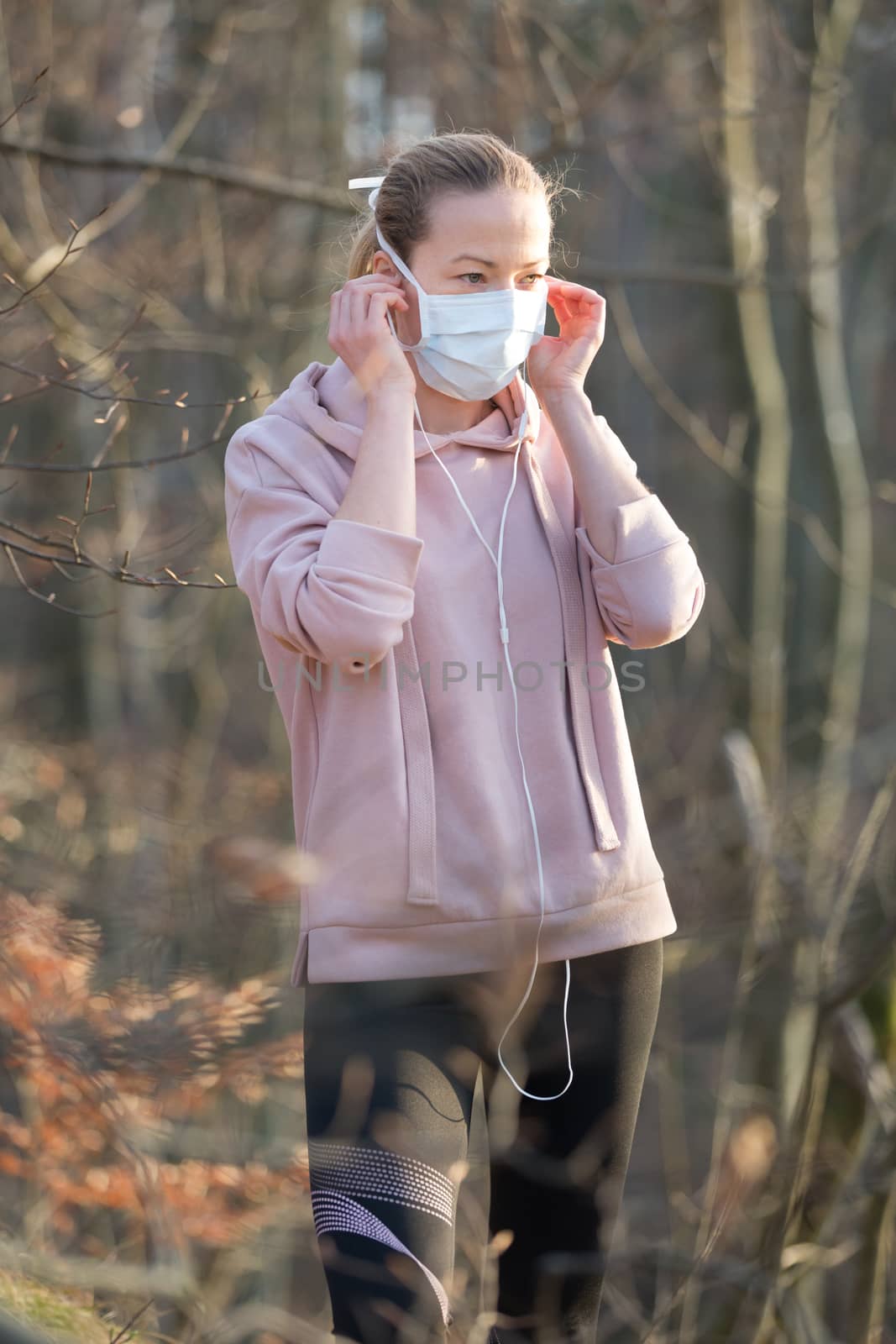 Portrait of caucasian sporty woman wearing medical protection face mask while walking in park, relaxing and listening to music. Corona virus, or Covid-19, is spreading all over the world.