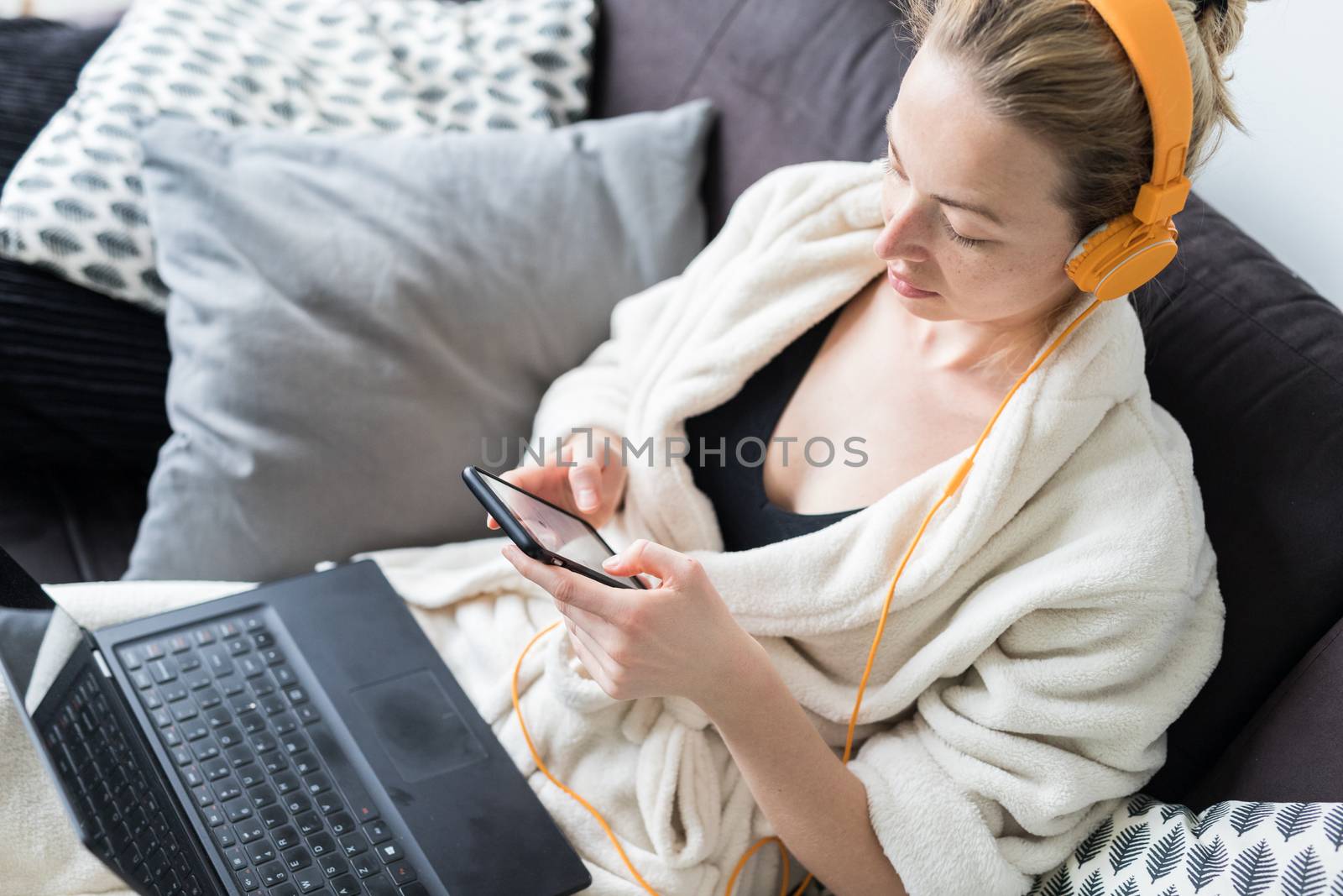 Woman in bathrobe being comfortable at her home sofa, using social media apps on phone for video chatting and stying connected with her loved ones. Social distancing. Stay at home.